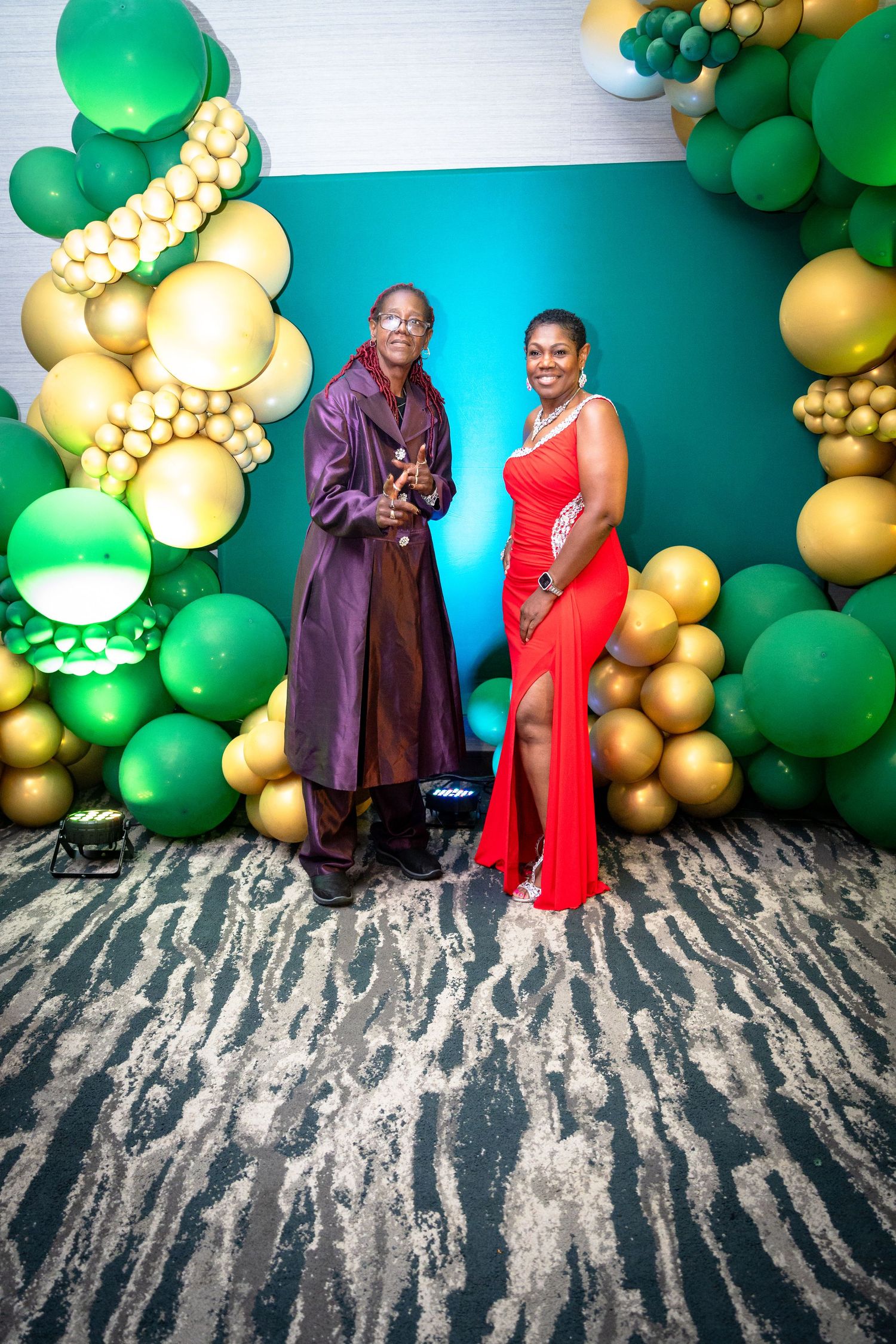 Festive party setup with elegant formal attire against turquoise backdrop decorated with green and gold balloon garlands.