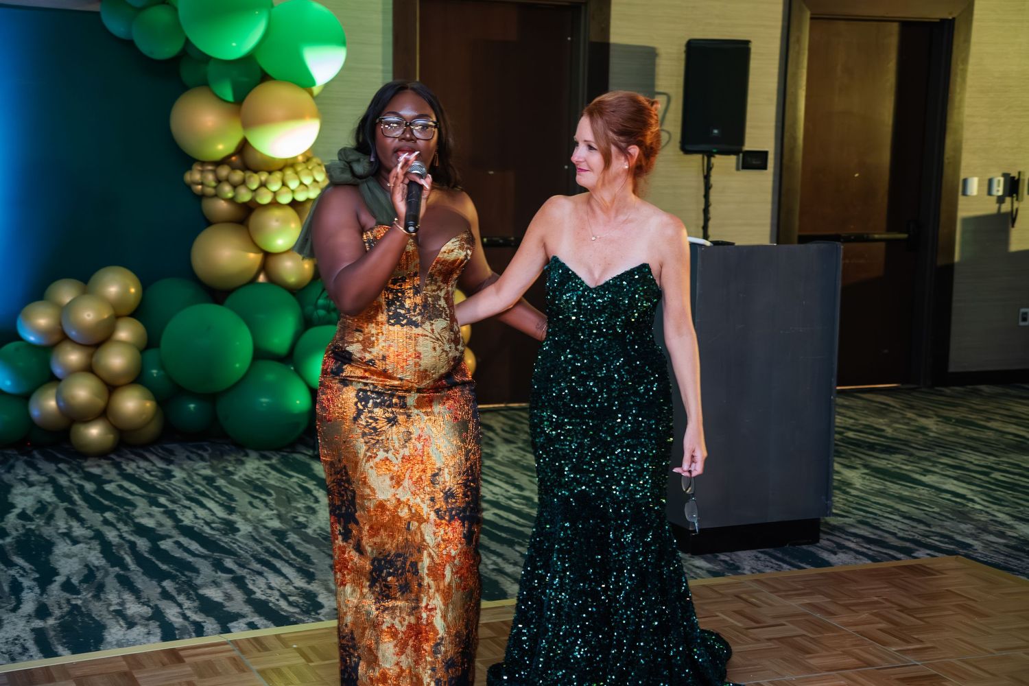 Two people in elegant dresses stand together at a formal event with gold balloon decorations in the background.