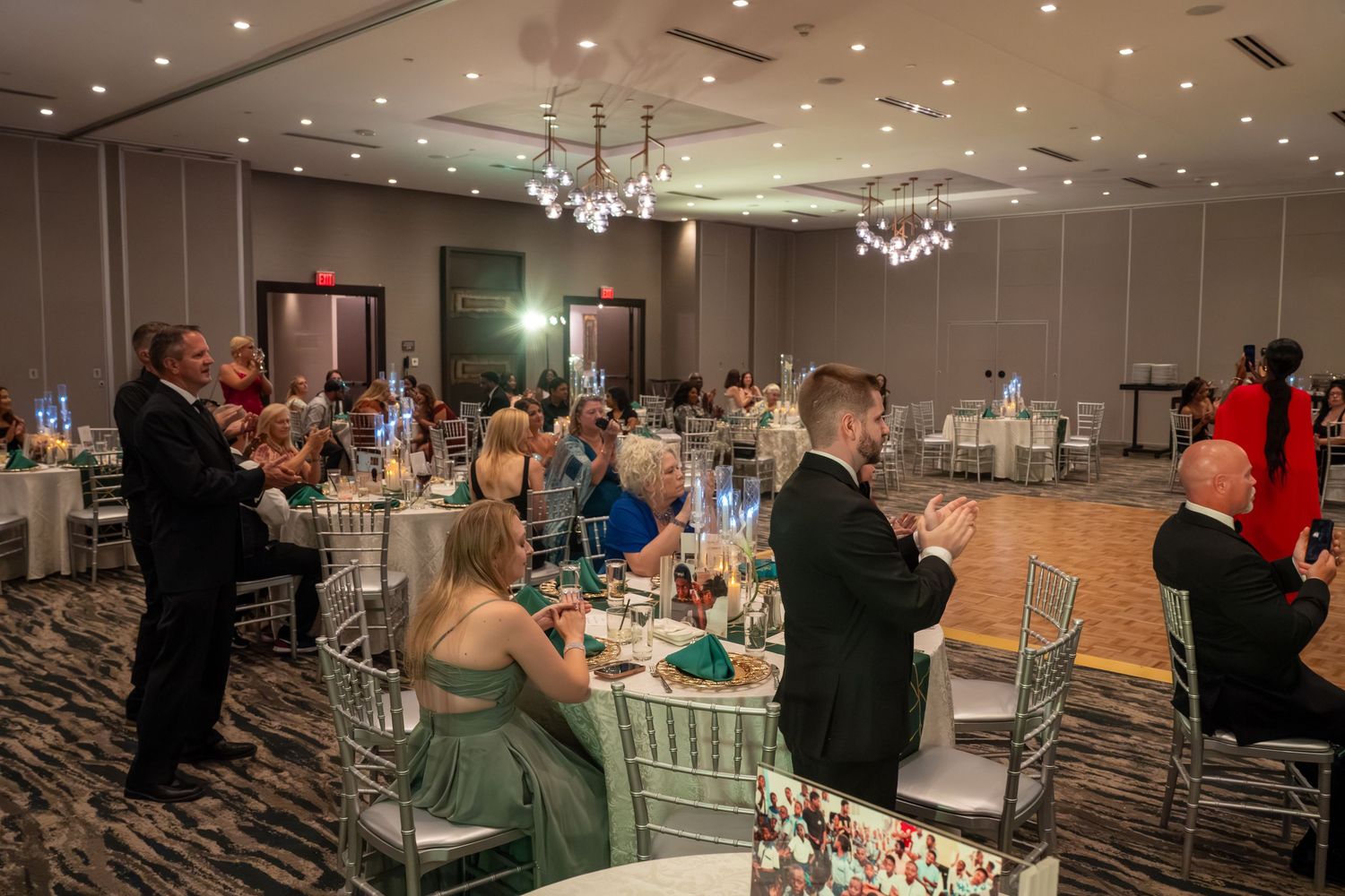 Wedding reception hall with crystal chandeliers, white chairs and guests seated at decorated tables.