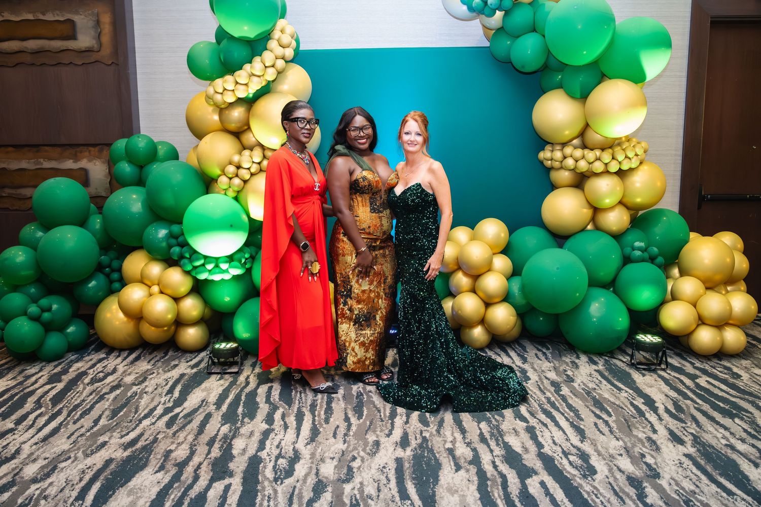 Three people pose in formal evening gowns against a backdrop of green and gold balloon decorations at an event.
