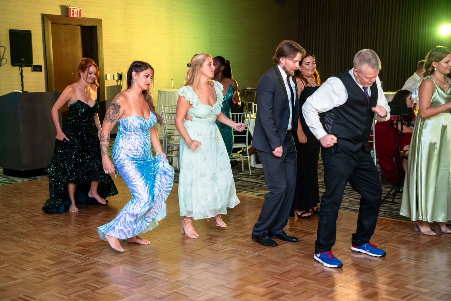 People dancing in a line at a wedding reception with colorful lighting and formal attire.