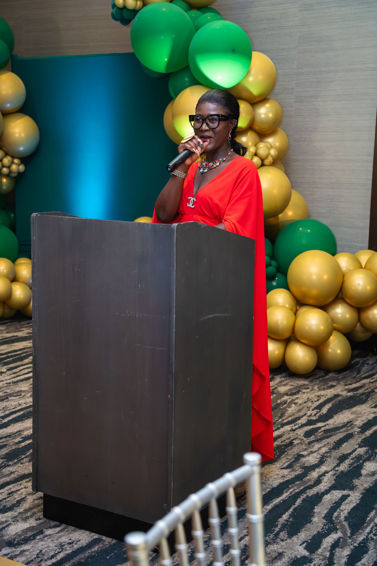 Speaker in a red dress stands at a podium against a backdrop of green and gold balloons at an event.