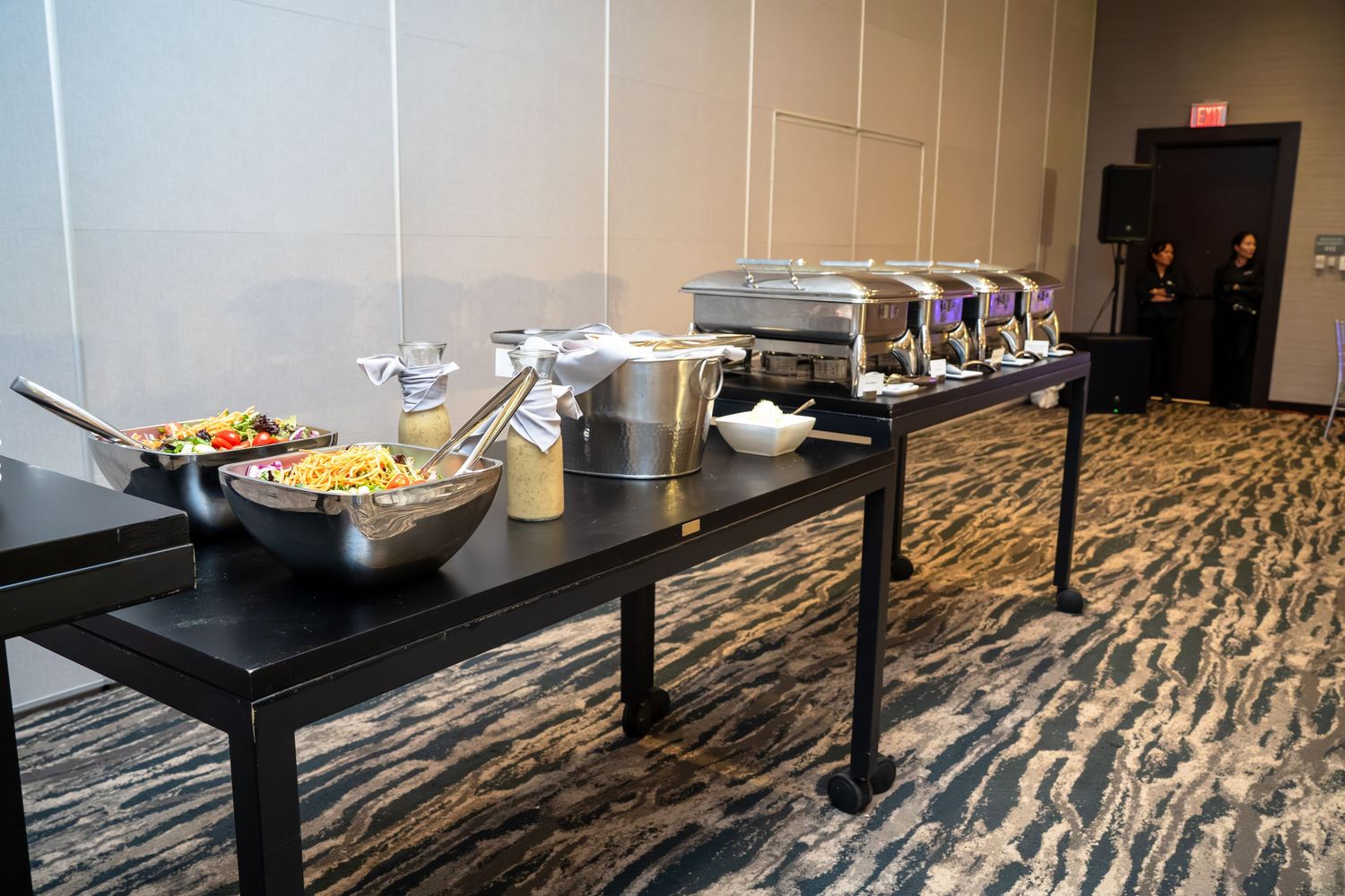 A modern conference room with black tables arranged in rows features a patterned carpet floor and buffet setup.
