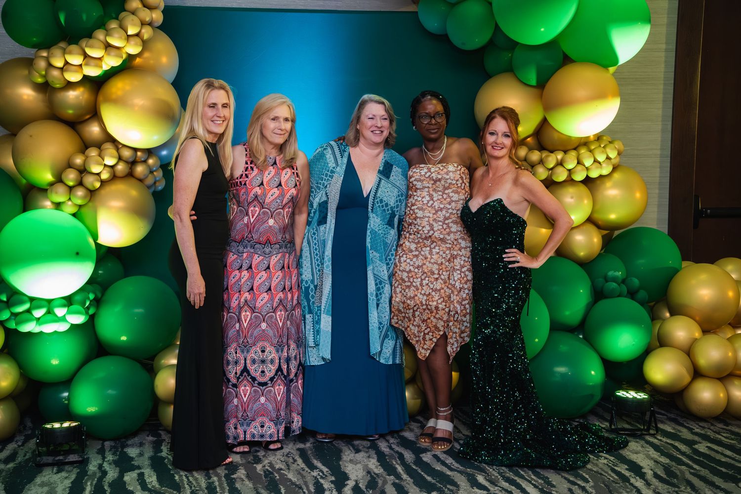 A group of five women pose together in formal dresses against a green and gold balloon arch backdrop at an event.