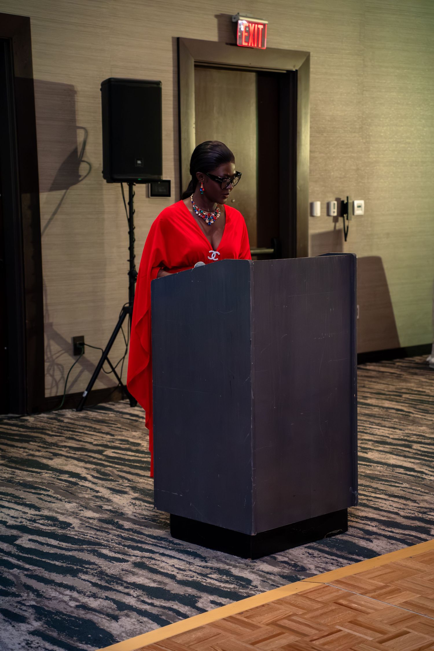 A speaker stands at a podium in a carpeted conference room with an exit sign visible in the background.