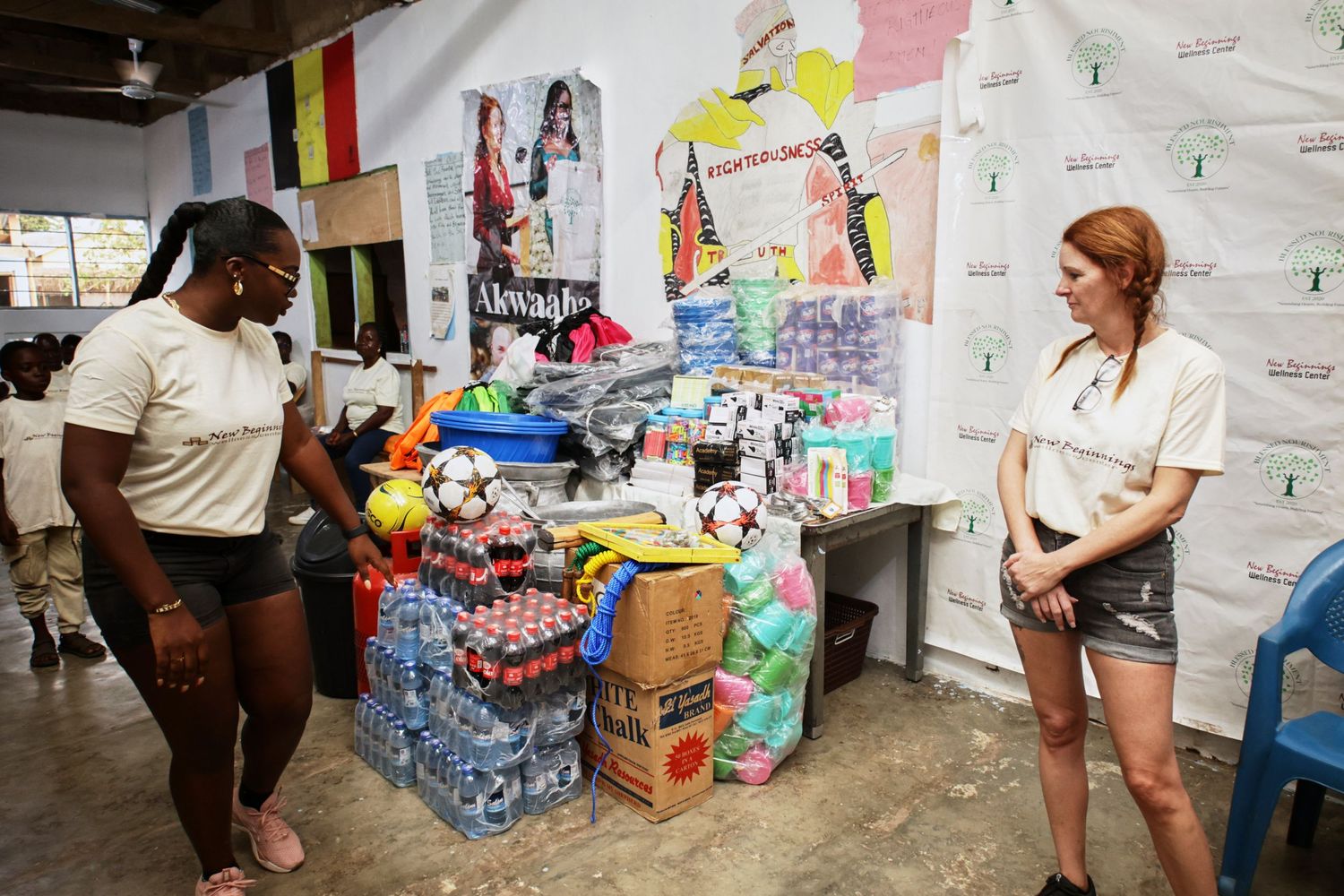 Two people chat next to stacked supplies and donations in a community space with decorated walls.
