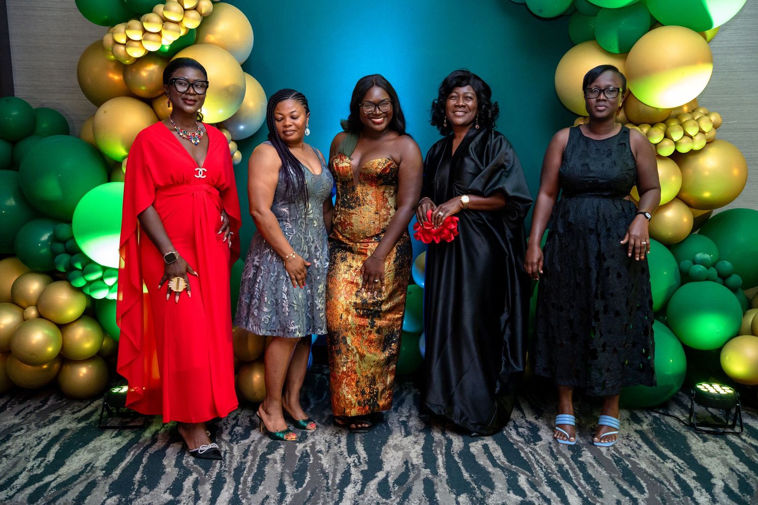Group photo of five women in formal attire standing against a festive backdrop with green and gold balloons.