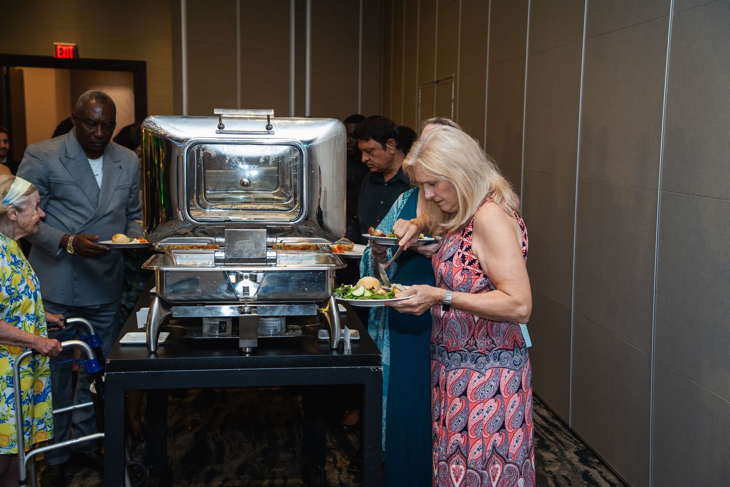 People gather around a buffet line with serving trays and dishes at what appears to be a catered event.