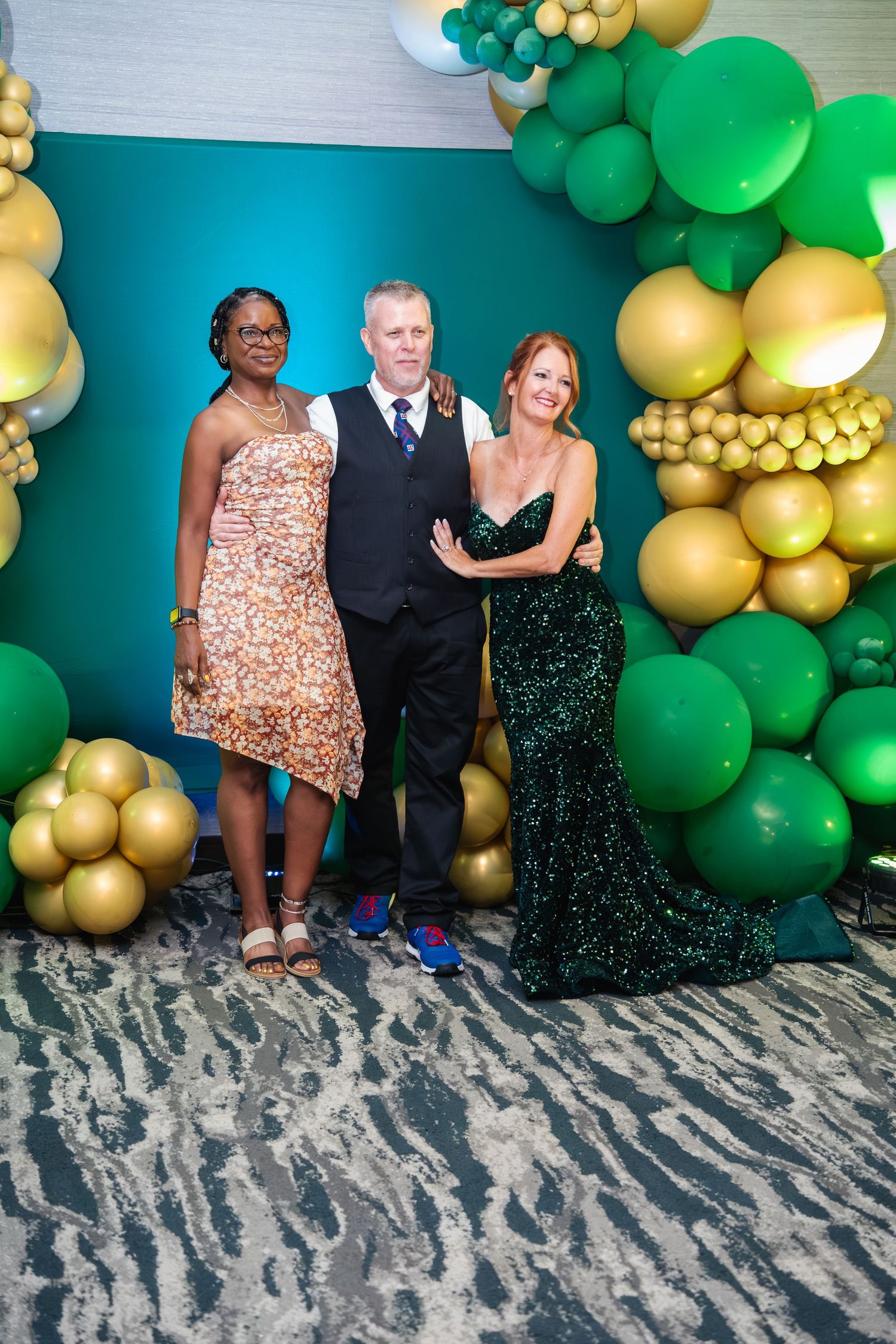 A festive party backdrop with green and gold balloon garland decorations against a blue wall.
