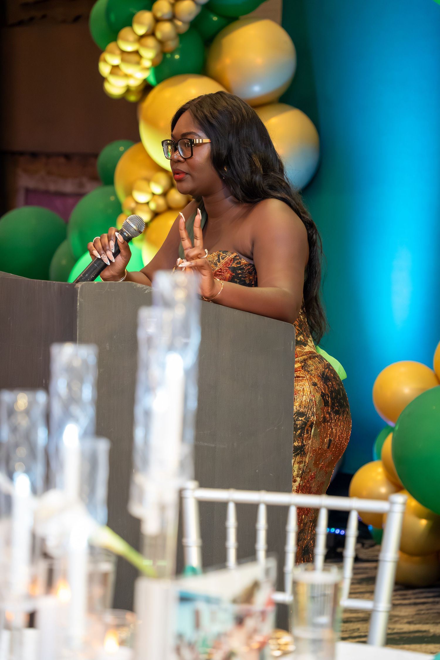 A speaker in a sparkly gold dress addresses an audience at an event decorated with green and gold balloons.