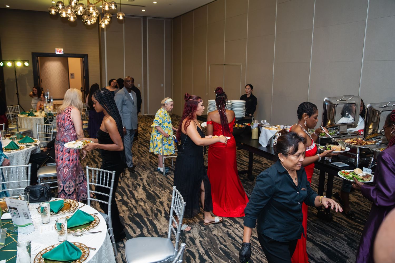 Guests gather around buffet tables at an elegant banquet with crystal chandeliers and green table settings.