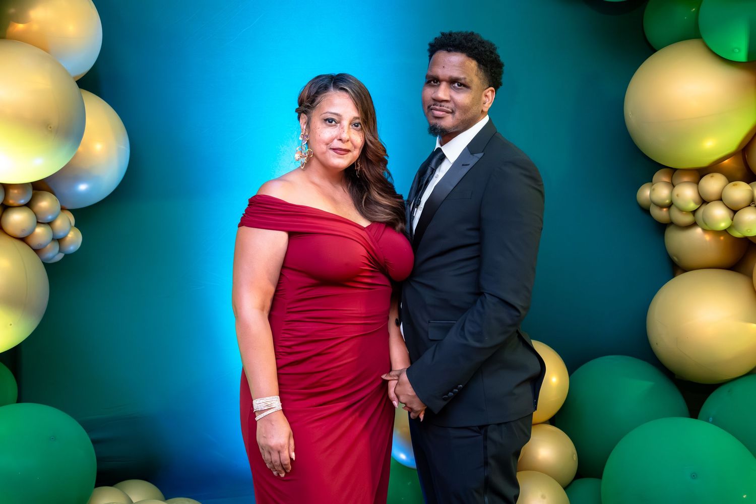 Couple in formal attire posing against a backdrop with gold and green balloon decorations.