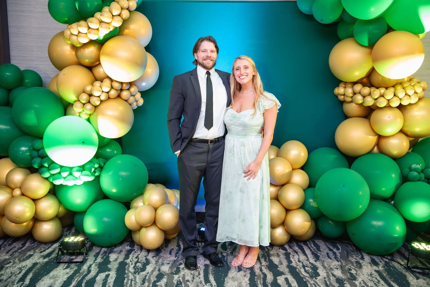 Wedding couple poses in front of elegant green and gold balloon arch decorations at their celebration.
