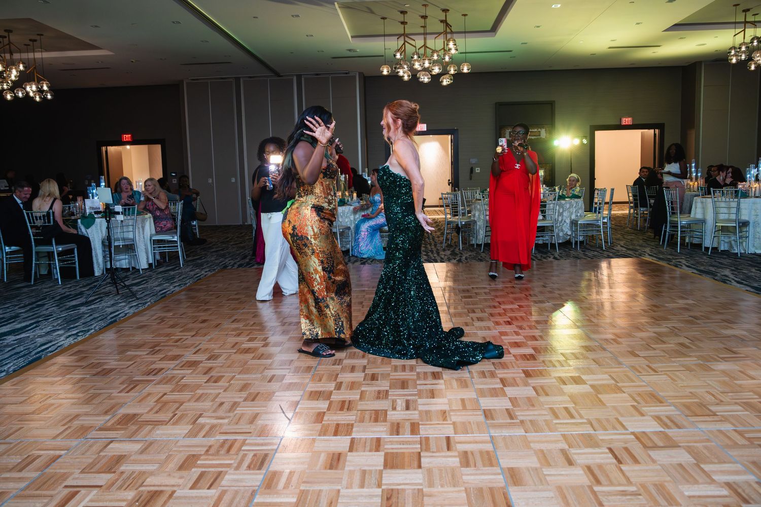 Dancers in formal attire move across a parquet dance floor at an elegant event venue with chandeliers and table settings.