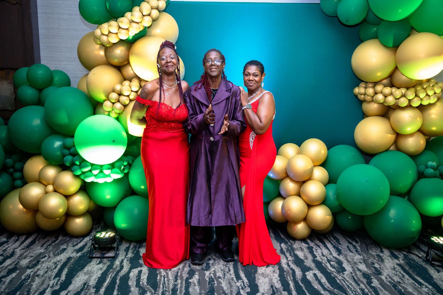 People pose against a teal wall decorated with green and gold balloon garlands for a festive celebration.