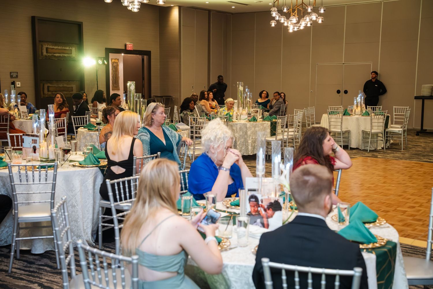 Wedding reception guests seated at round tables with white linens in an elegant banquet hall with crystal chandeliers.