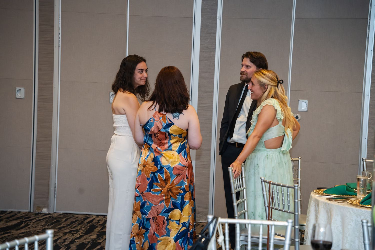 A group of women in formal attire chat and mingle at an indoor event with gray walls and table settings.