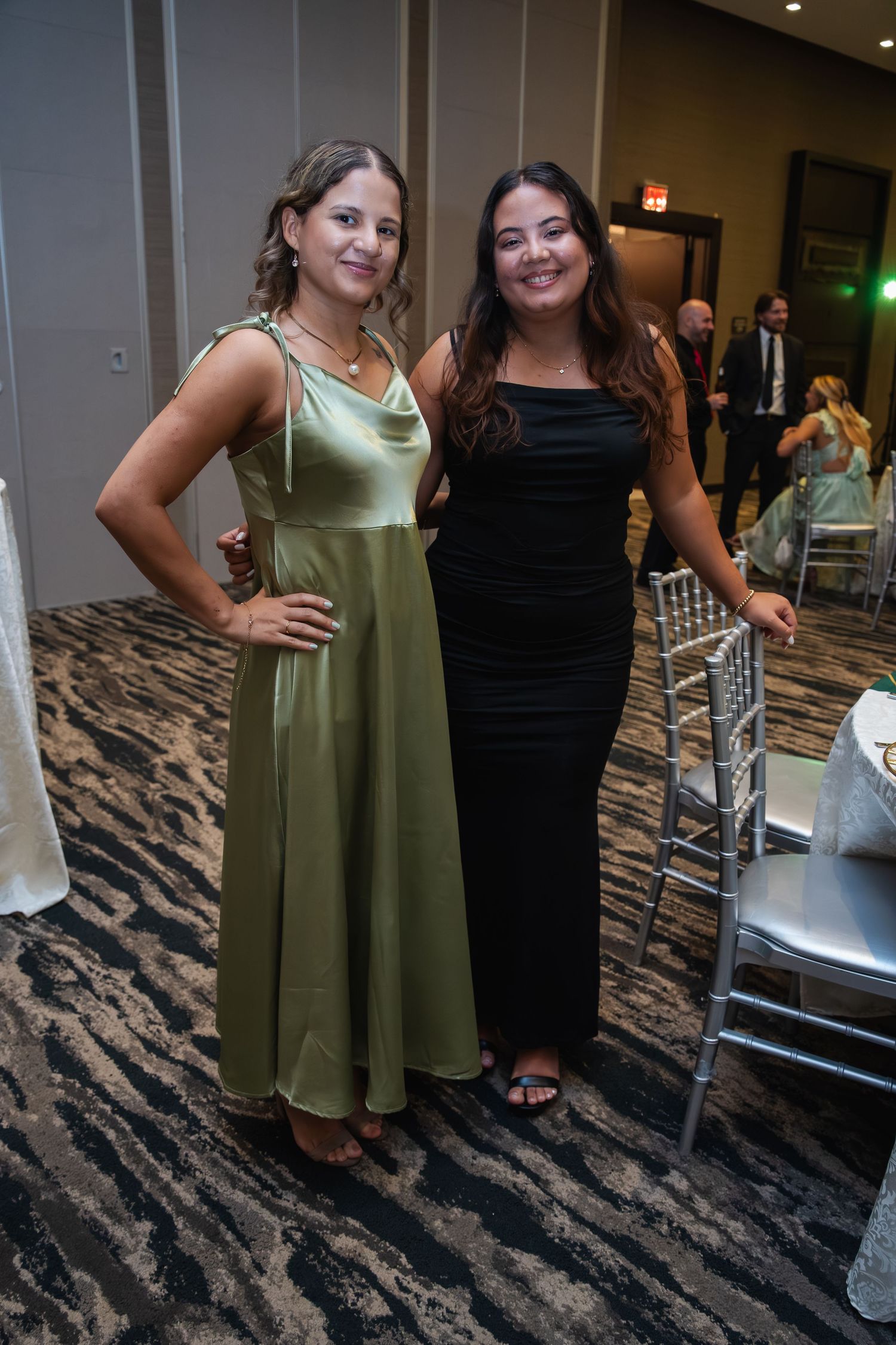 Two women pose together at an event wearing contrasting black and sage green dresses on a patterned carpet floor.