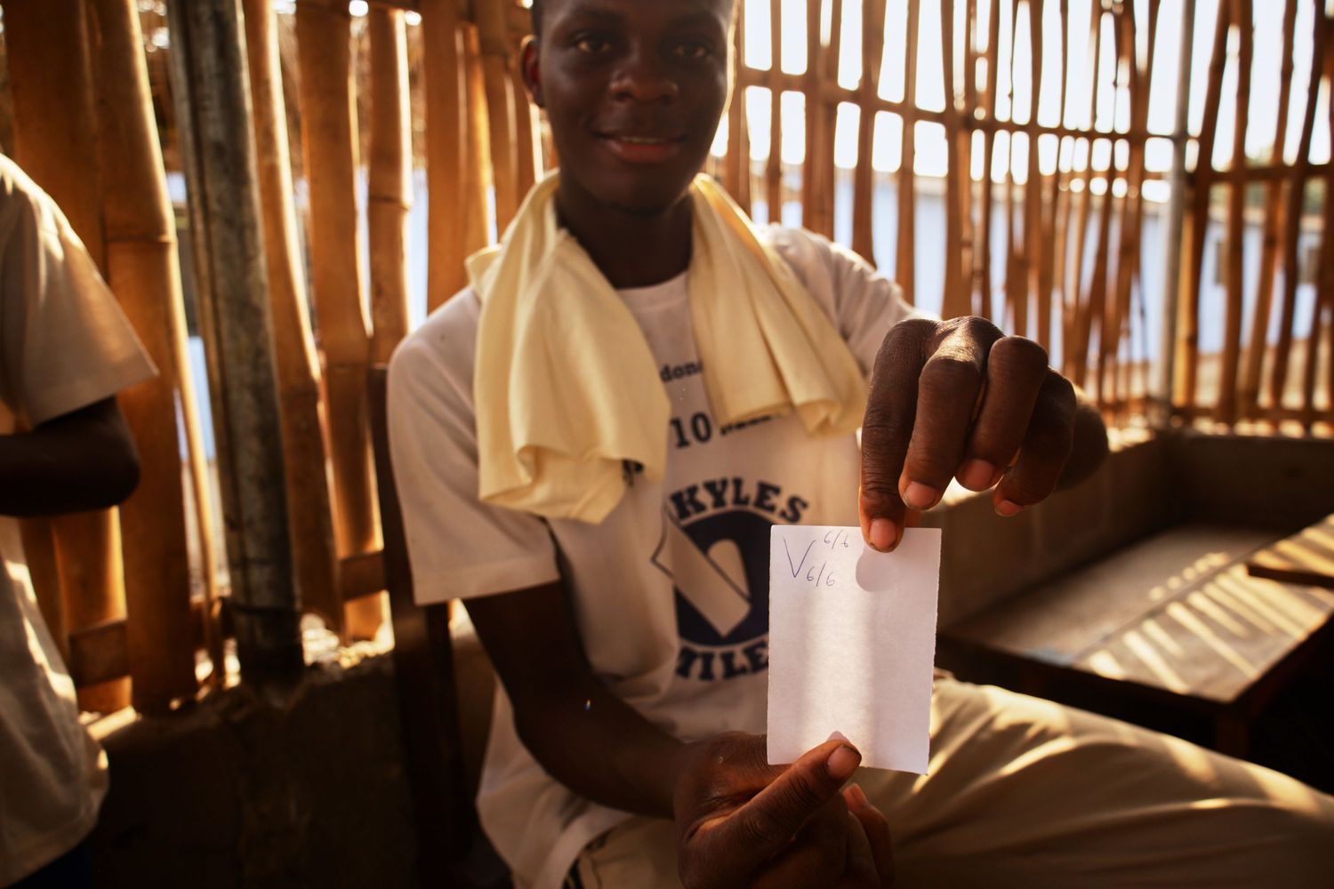 Person in white shirt holds up small container in sunlit bamboo-walled classroom.