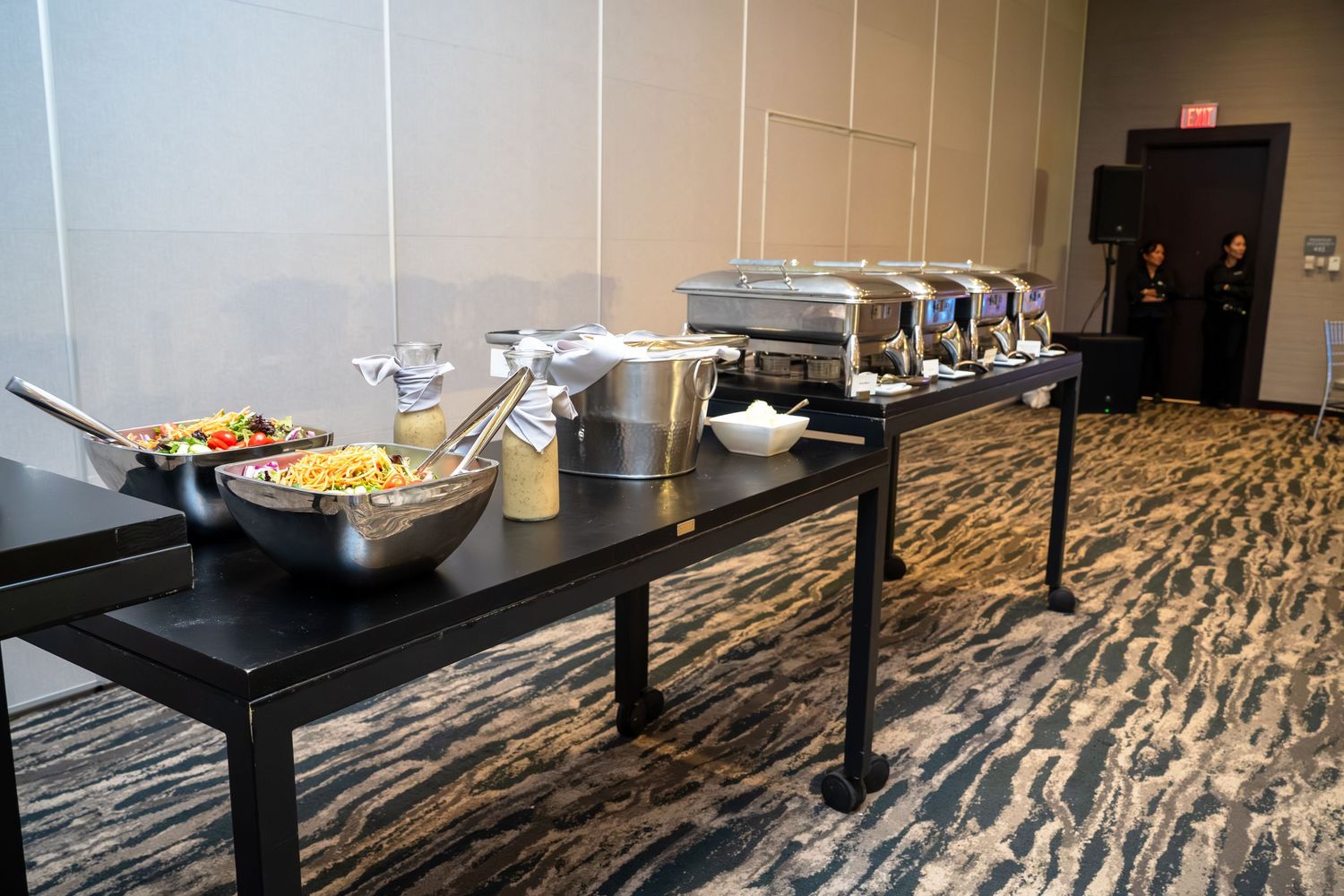 A modern conference room buffet setup with food warmers and serving stations on dark tables against patterned carpet.