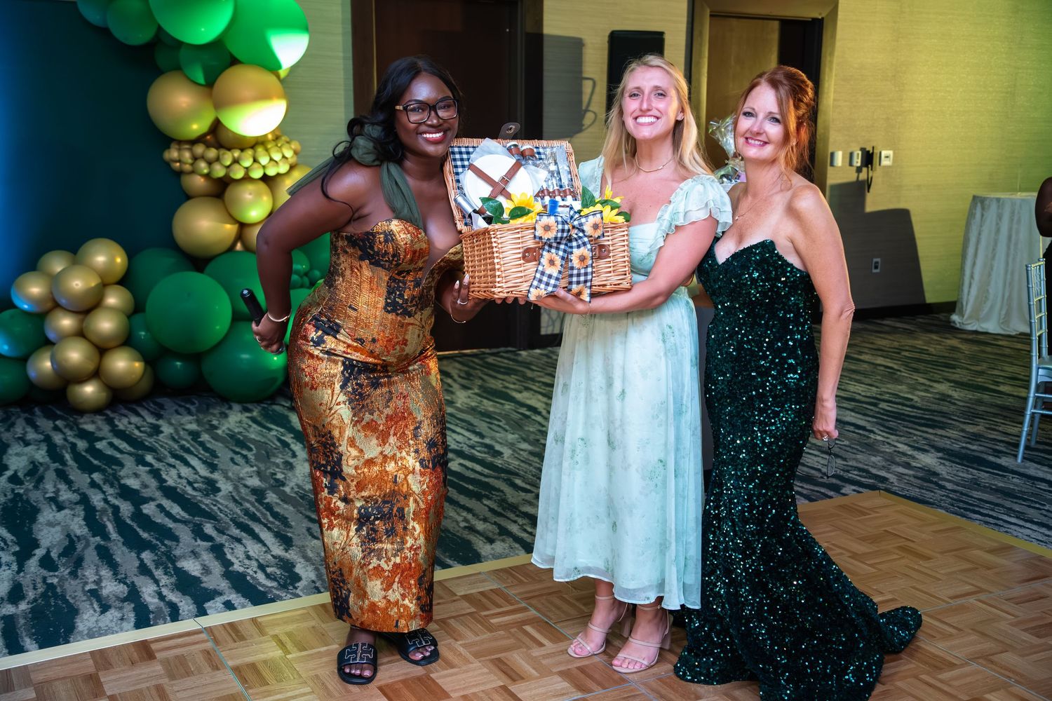 People dance and celebrate at a festive event with green and gold balloon decorations in an indoor venue.