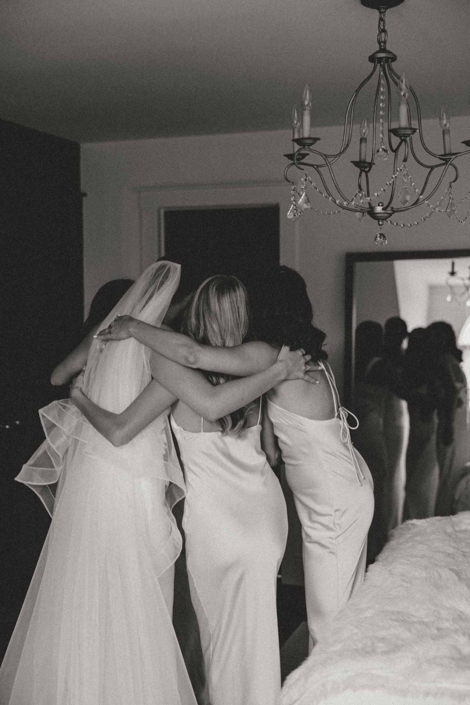 Black and white photo of bride and bridesmaids embracing from behind in a chandelier-lit room.