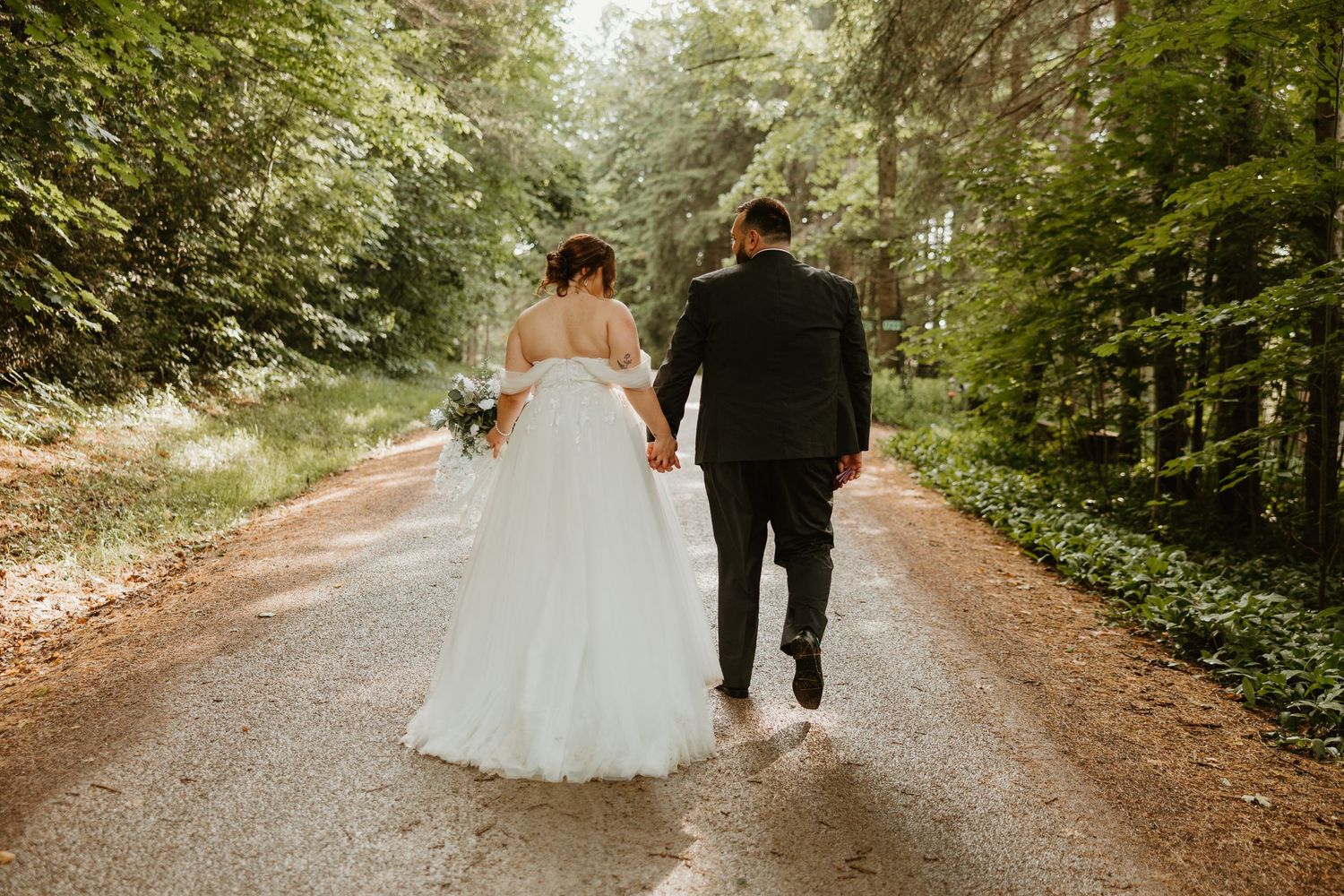 Bride and groom walking hand-in-hand down a sunlit forest path on their wedding day.