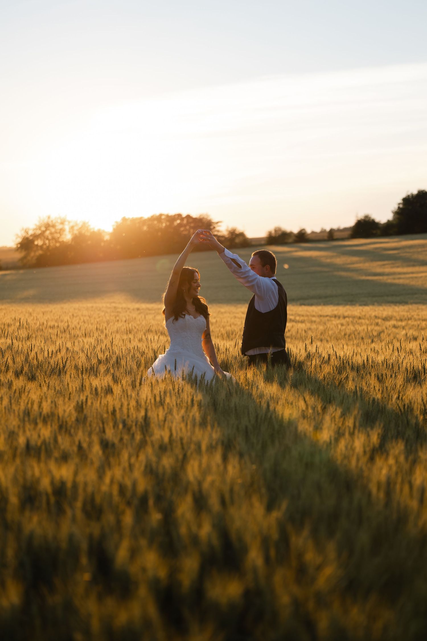 Bride and groom dancing at sunset in golden wheat field, silhouetted against warm glowing sky.