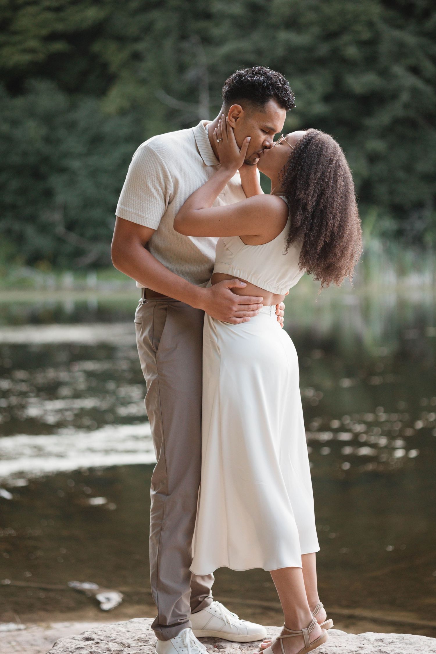 Couple sharing a romantic kiss by a peaceful lake surrounded by lush green trees.
