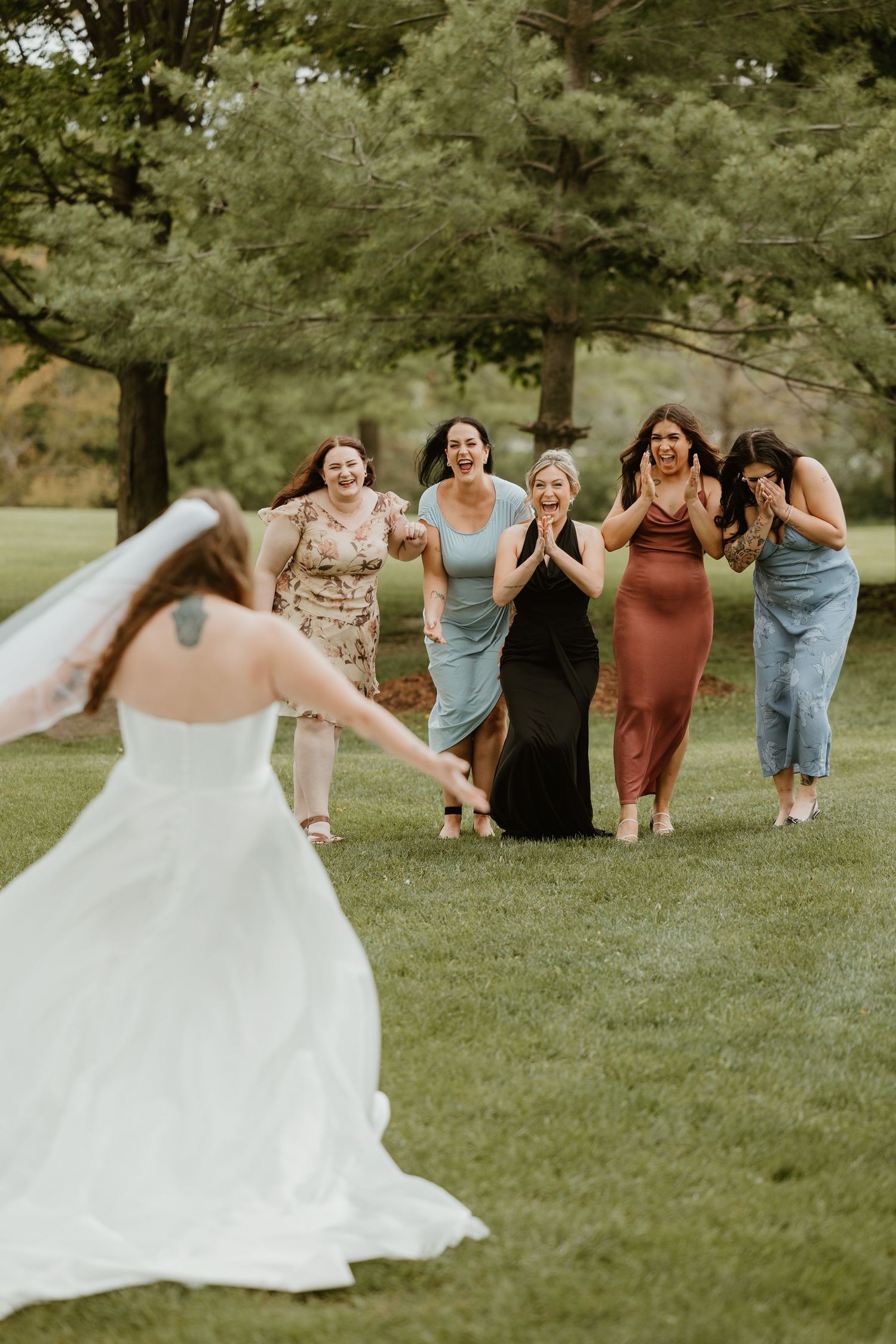 Bridesmaids react with joyful excitement during an outdoor first look reveal with the bride on grass.