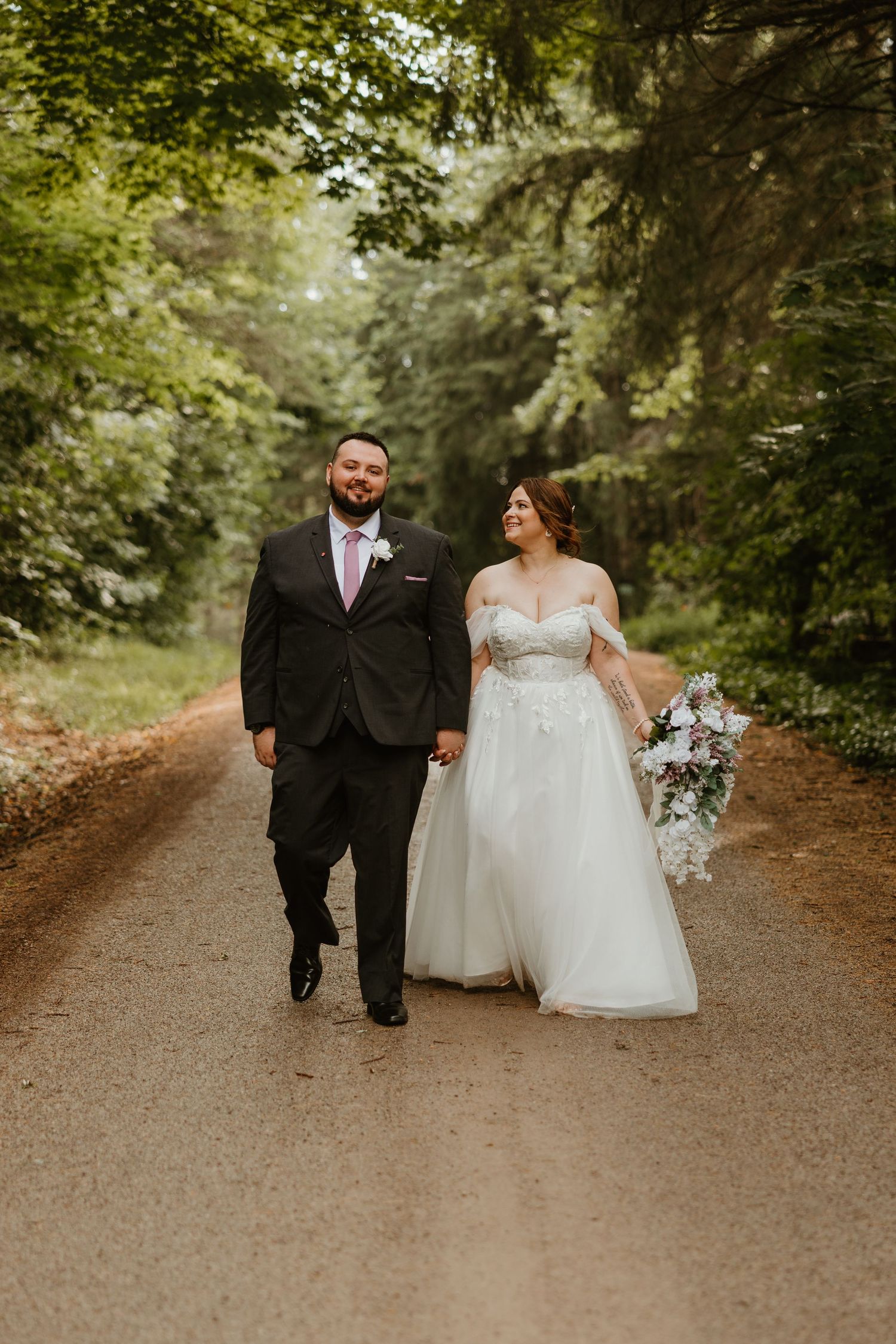 Bride in off-shoulder ballgown and groom in dark suit walk hand in hand down a tree-lined gravel path.