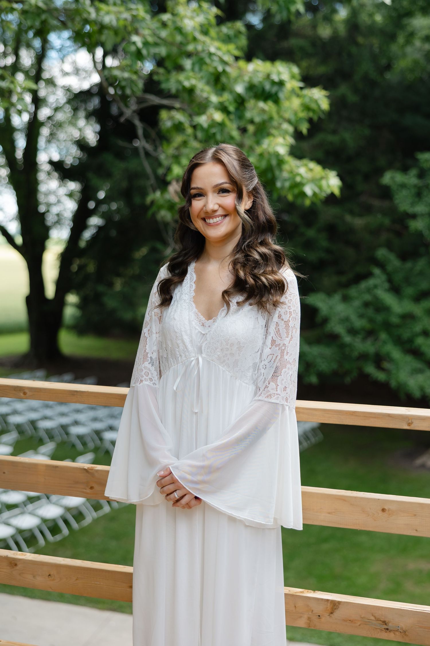 Smiling bride in white lace bell-sleeve gown standing on wooden deck with lush green trees backdrop.