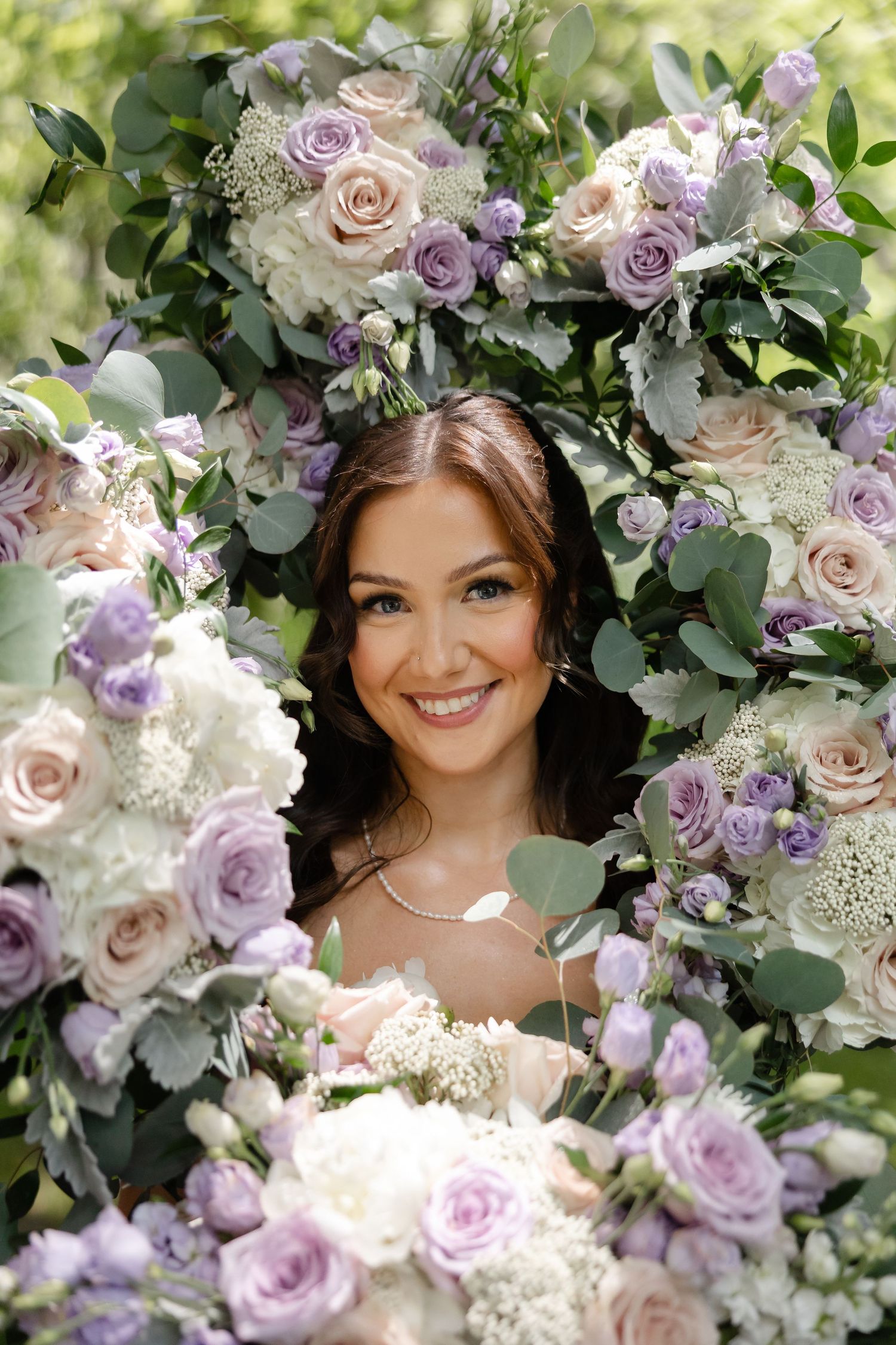 Smiling bride framed by lavender and blush roses, eucalyptus, and white floral arrangements in a lush garden setting.