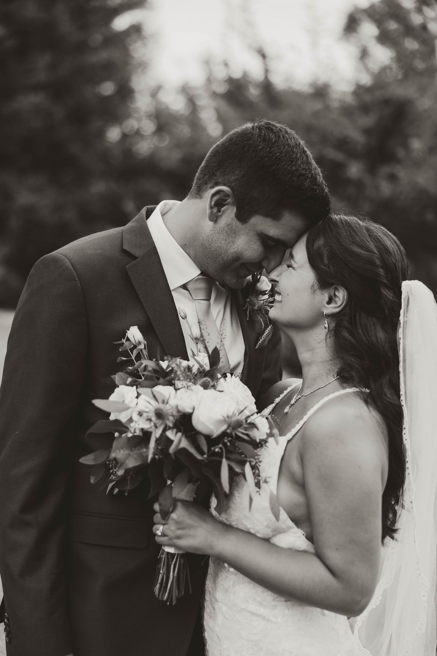 Black and white photo of bride and groom sharing a tender moment with a floral bouquet outdoors.