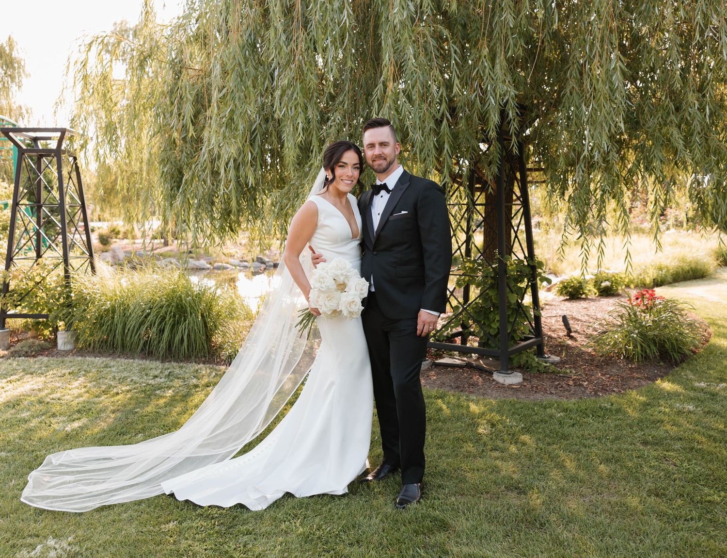 Bride and groom posing under a weeping willow tree in a lush garden setting.