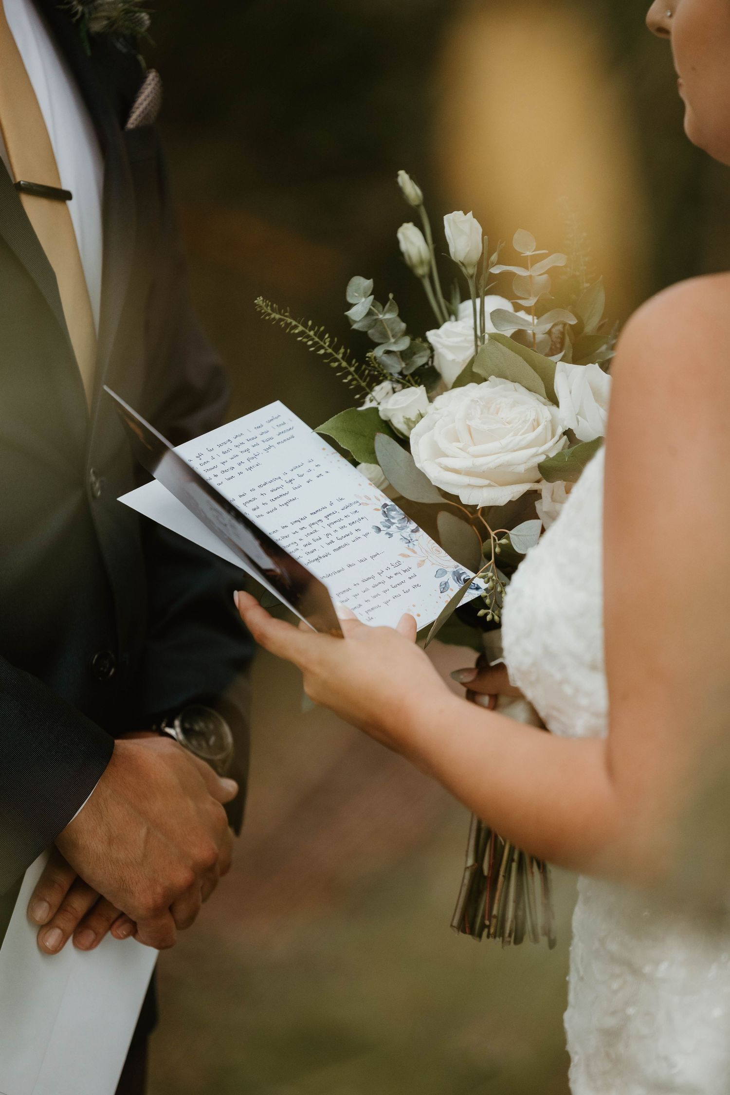 Bride reads handwritten vows while holding white rose bouquet during outdoor wedding ceremony.