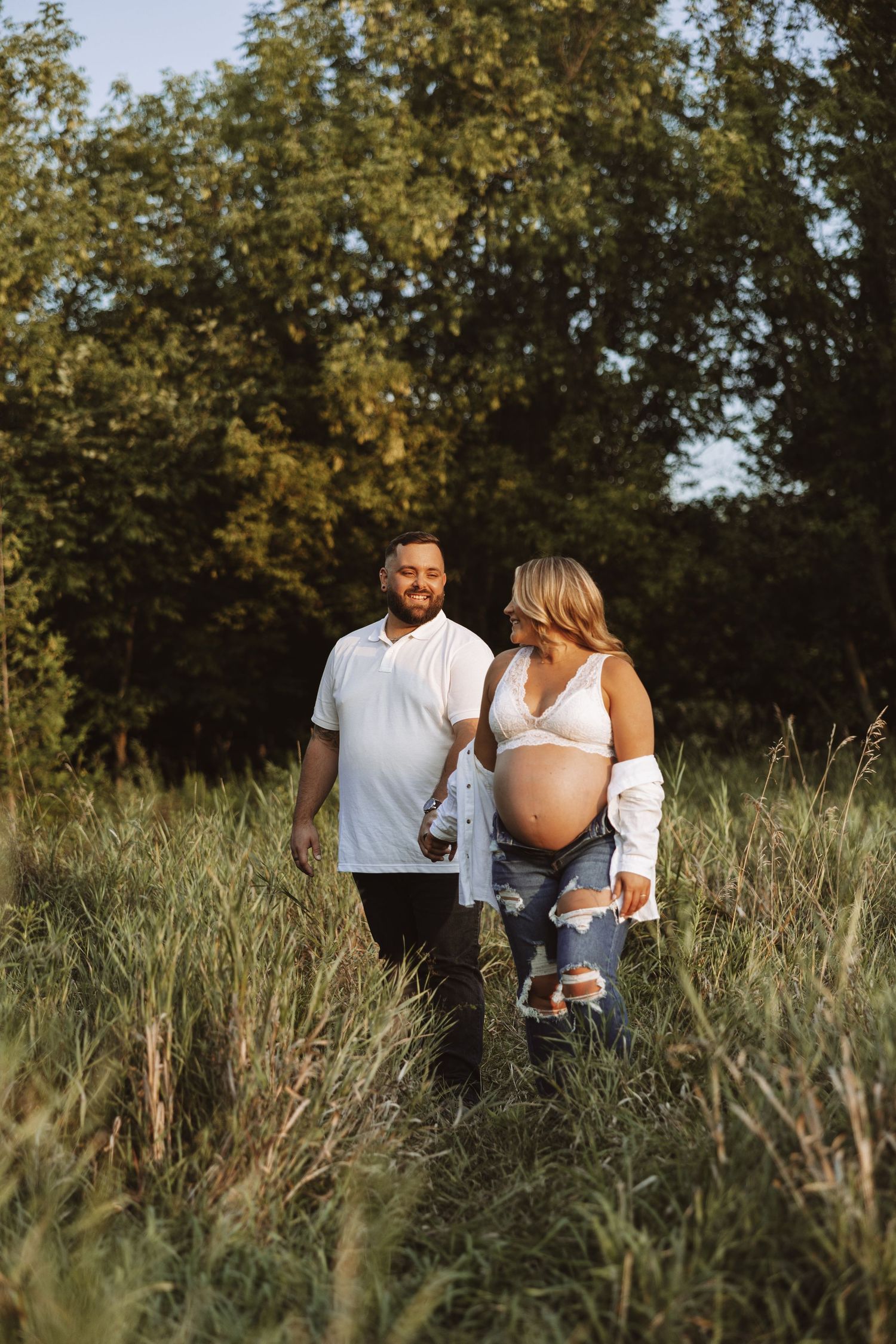 Pregnant couple walking through tall grass field in golden hour light.