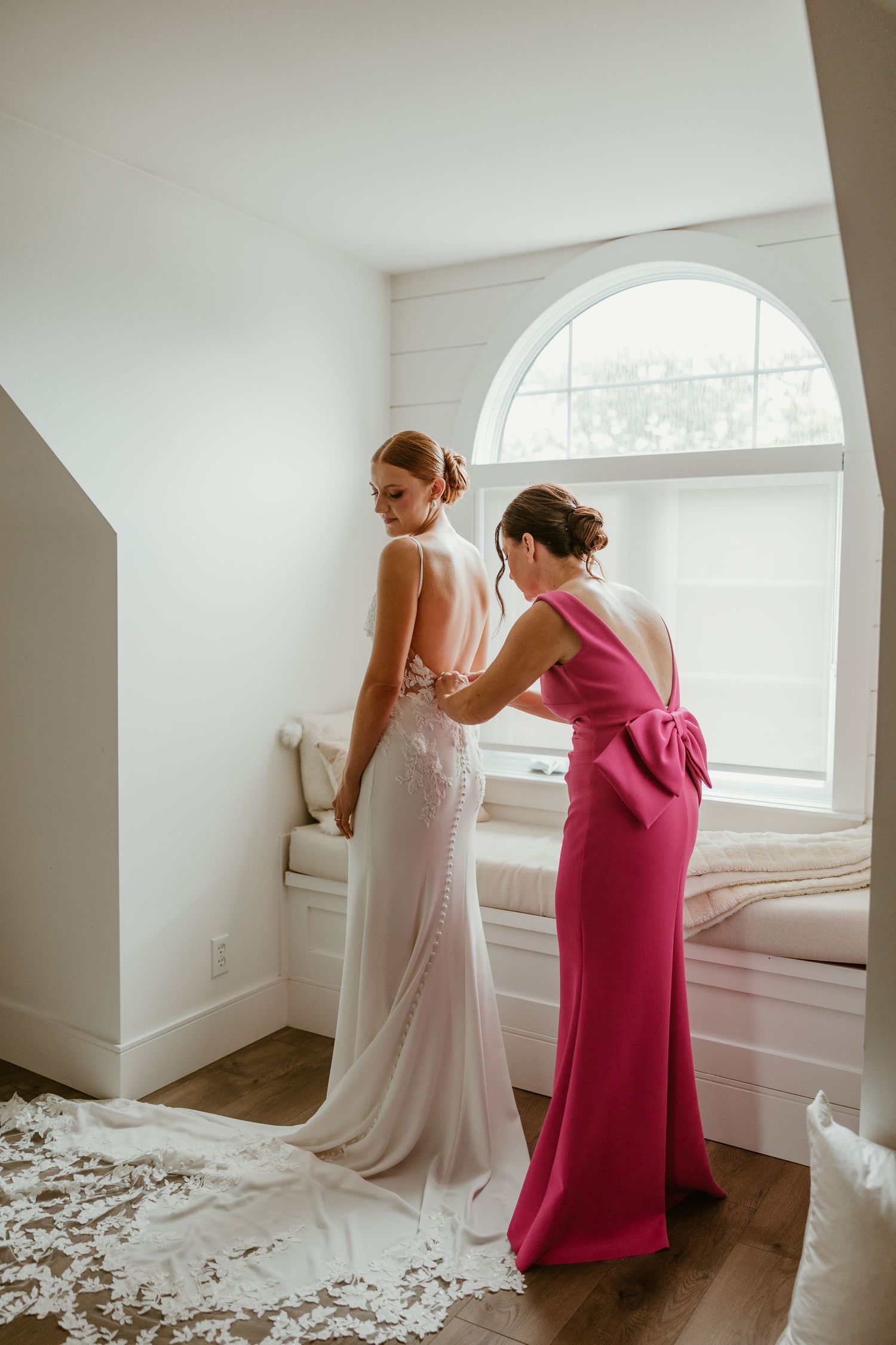 Bridesmaid in pink bow dress helping bride with her white lace wedding gown near an arched window.