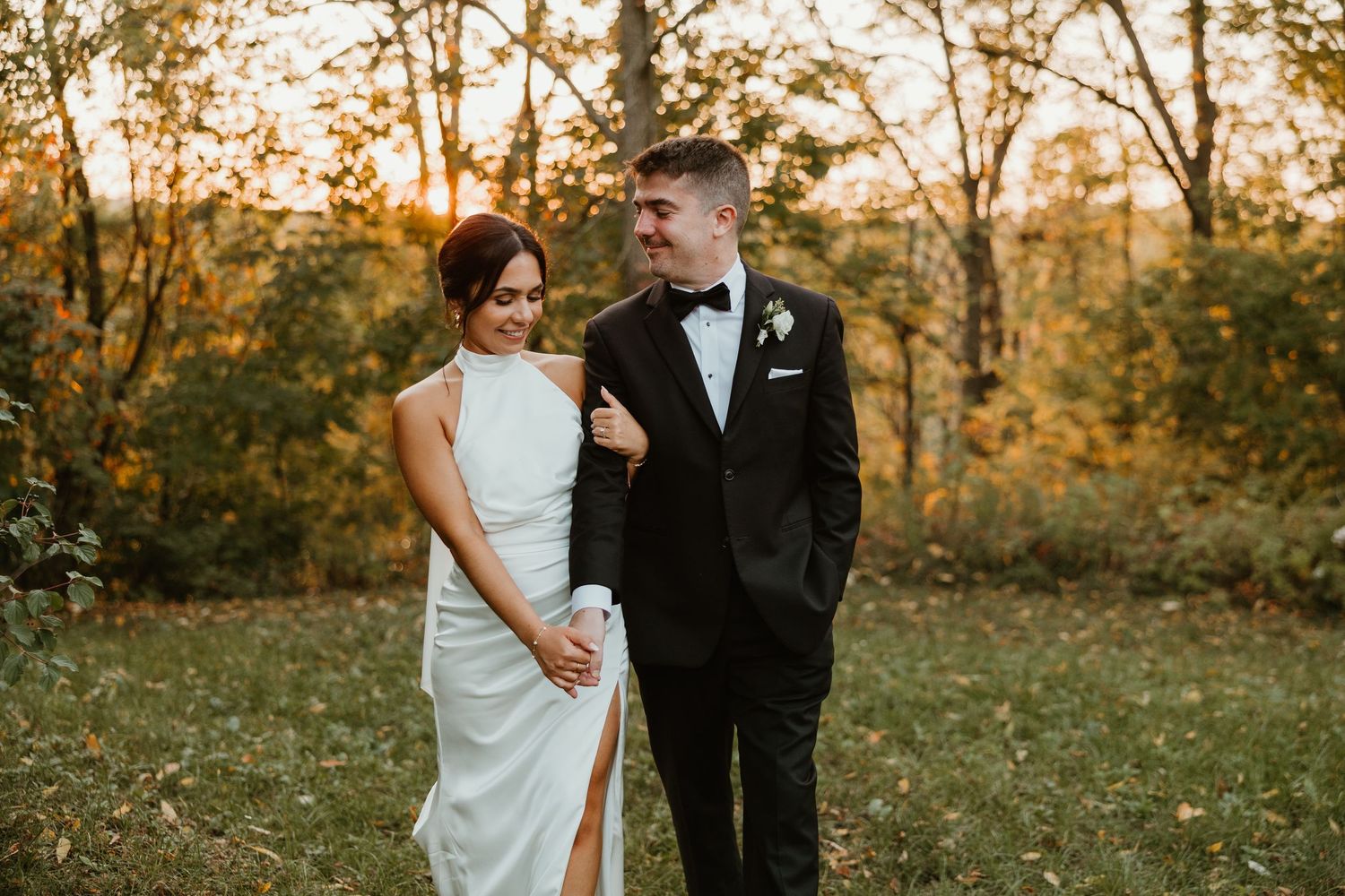 Bride in sleek white halter gown and groom in black tuxedo share a tender moment in a golden autumn forest setting.