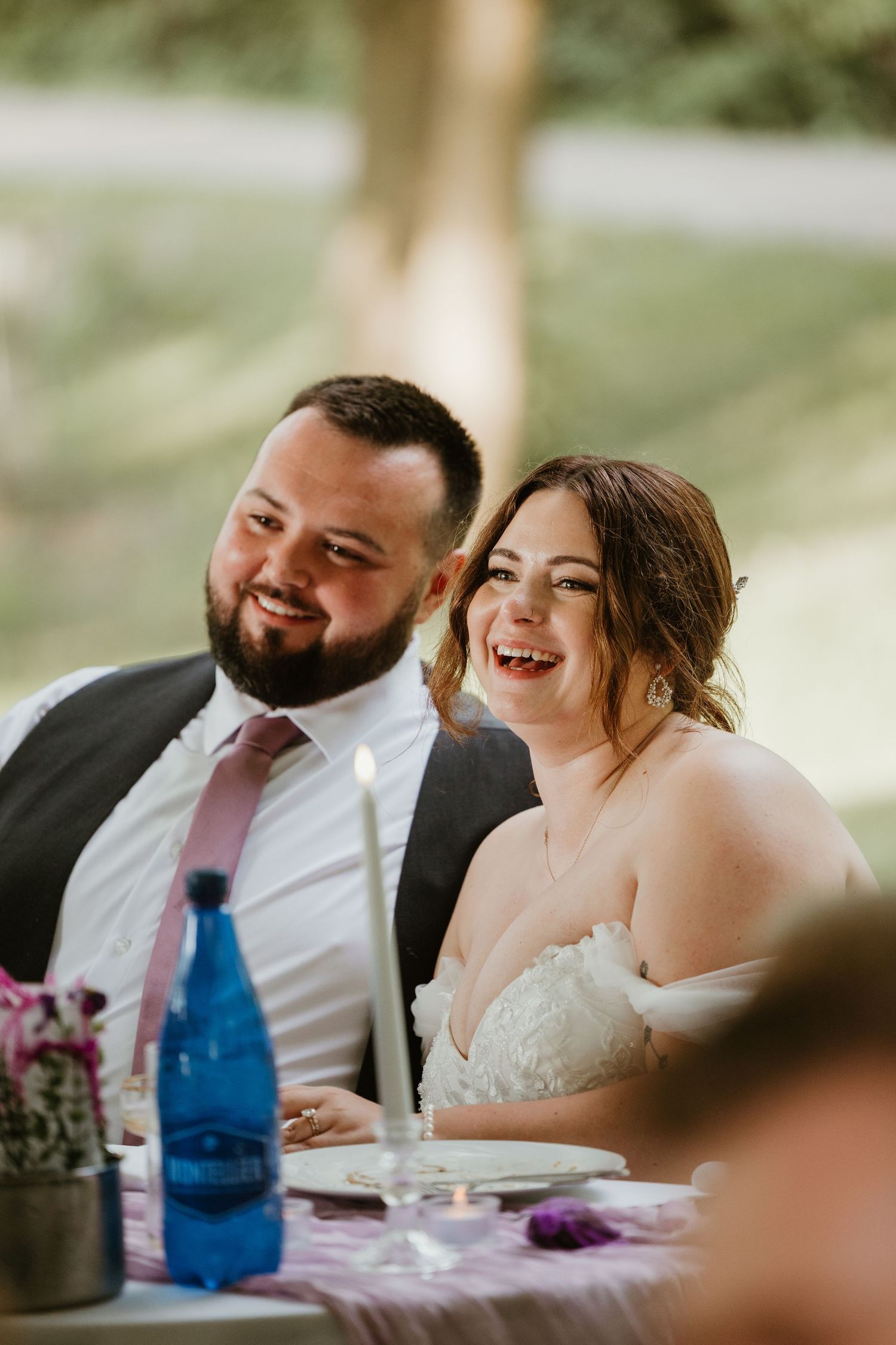 Smiling bride and bearded groom laughing together at outdoor wedding reception table.