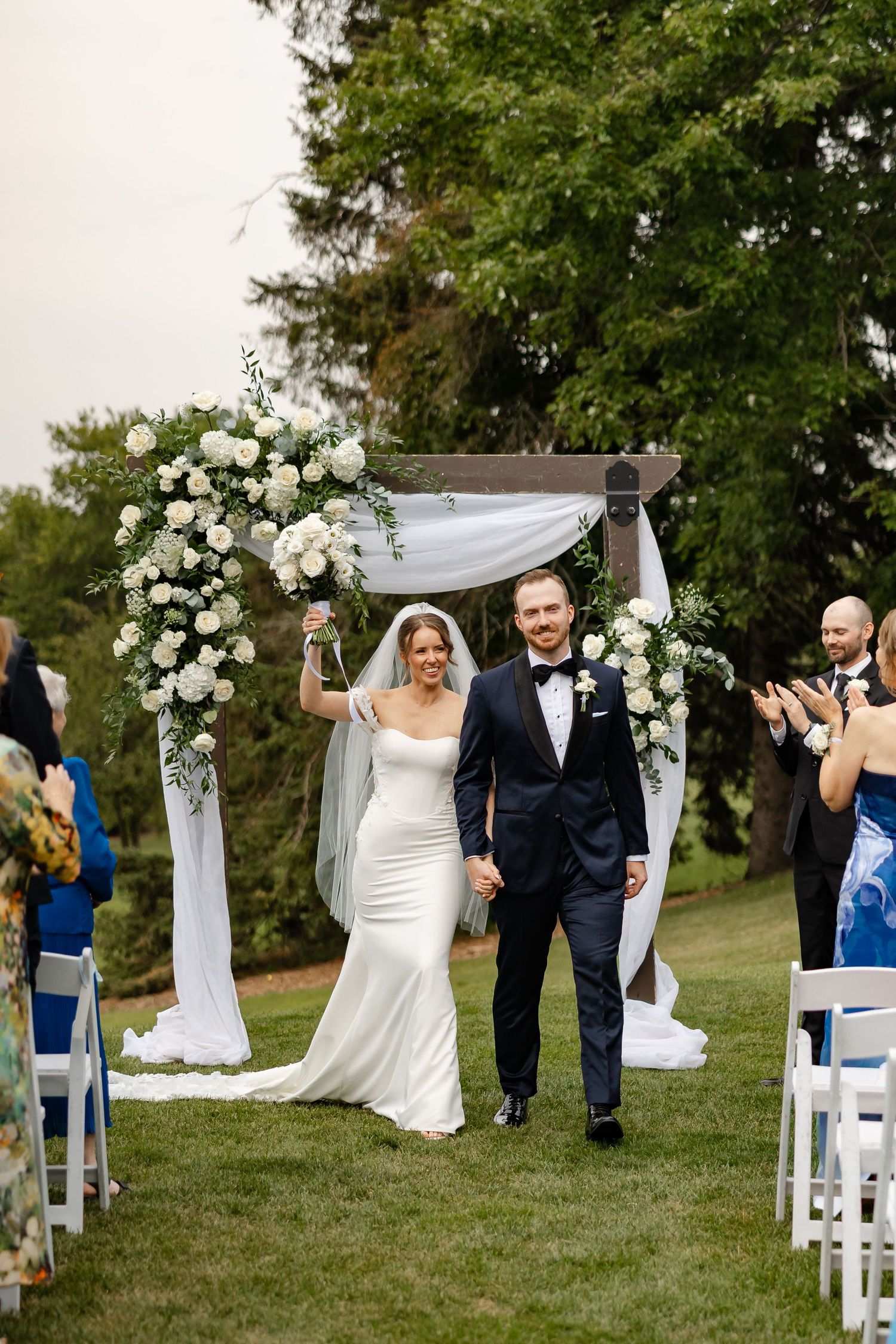 Bride and groom walk down outdoor aisle after wedding ceremony under floral arch with white roses and greenery.