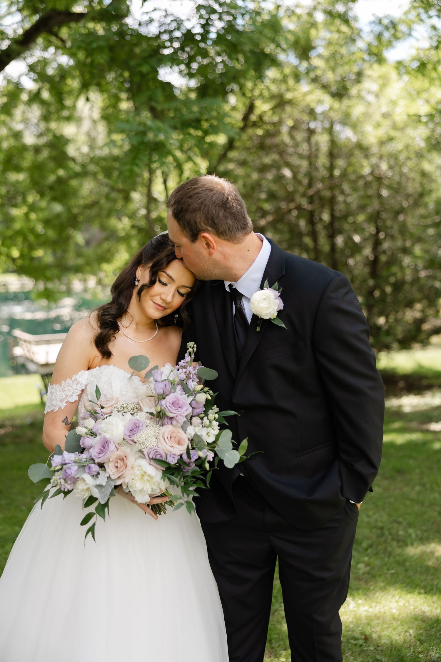 Groom tenderly kisses bride on the head as she holds lavender and white floral bouquet outdoors.