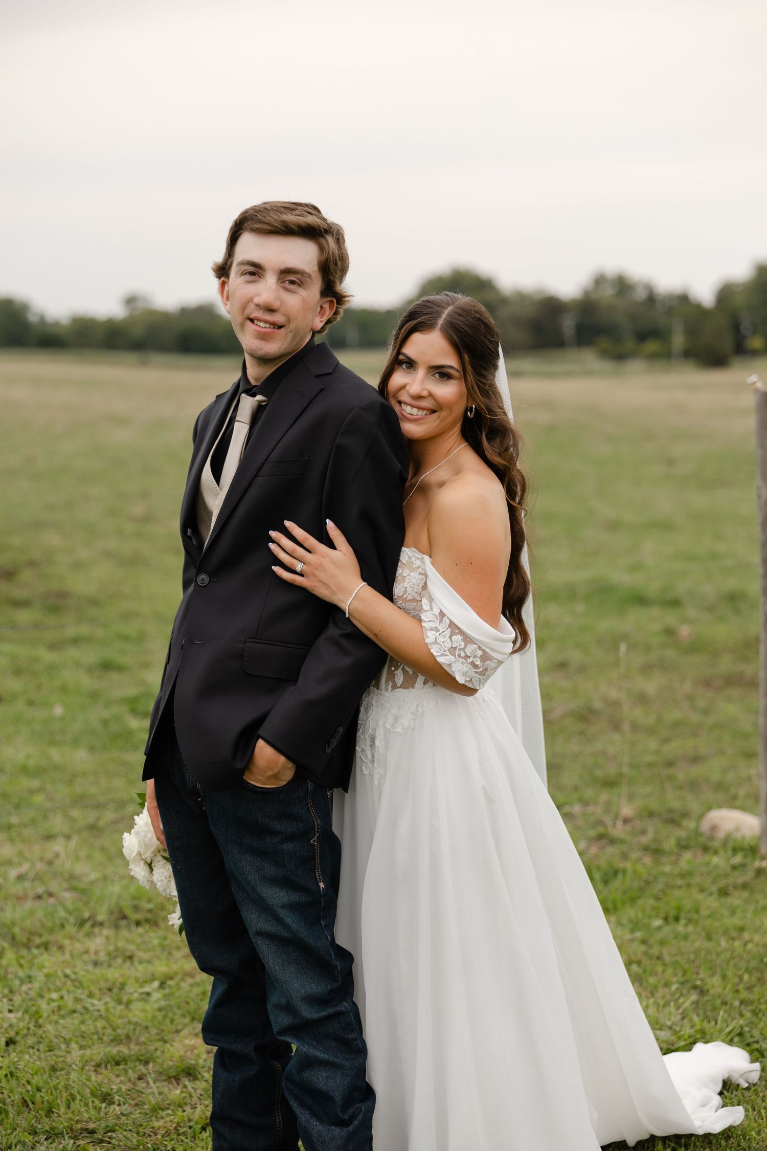 Smiling bride in an off-the-shoulder white lace gown and veil embraces a groom in a dark jacket and jeans in a grassy field.
