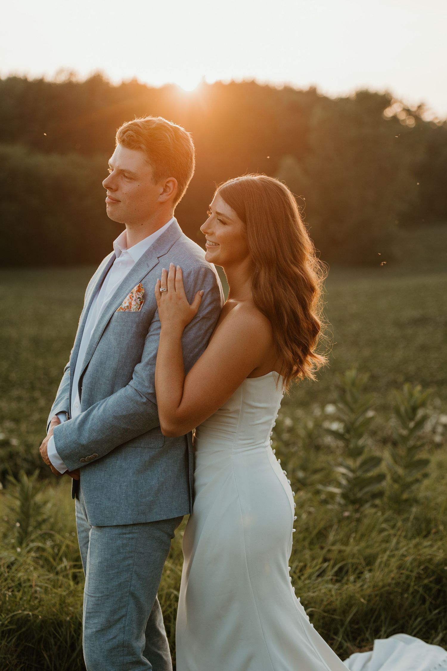 Bride and groom standing back to back in a field at golden hour sunset, bride in sleek white gown.