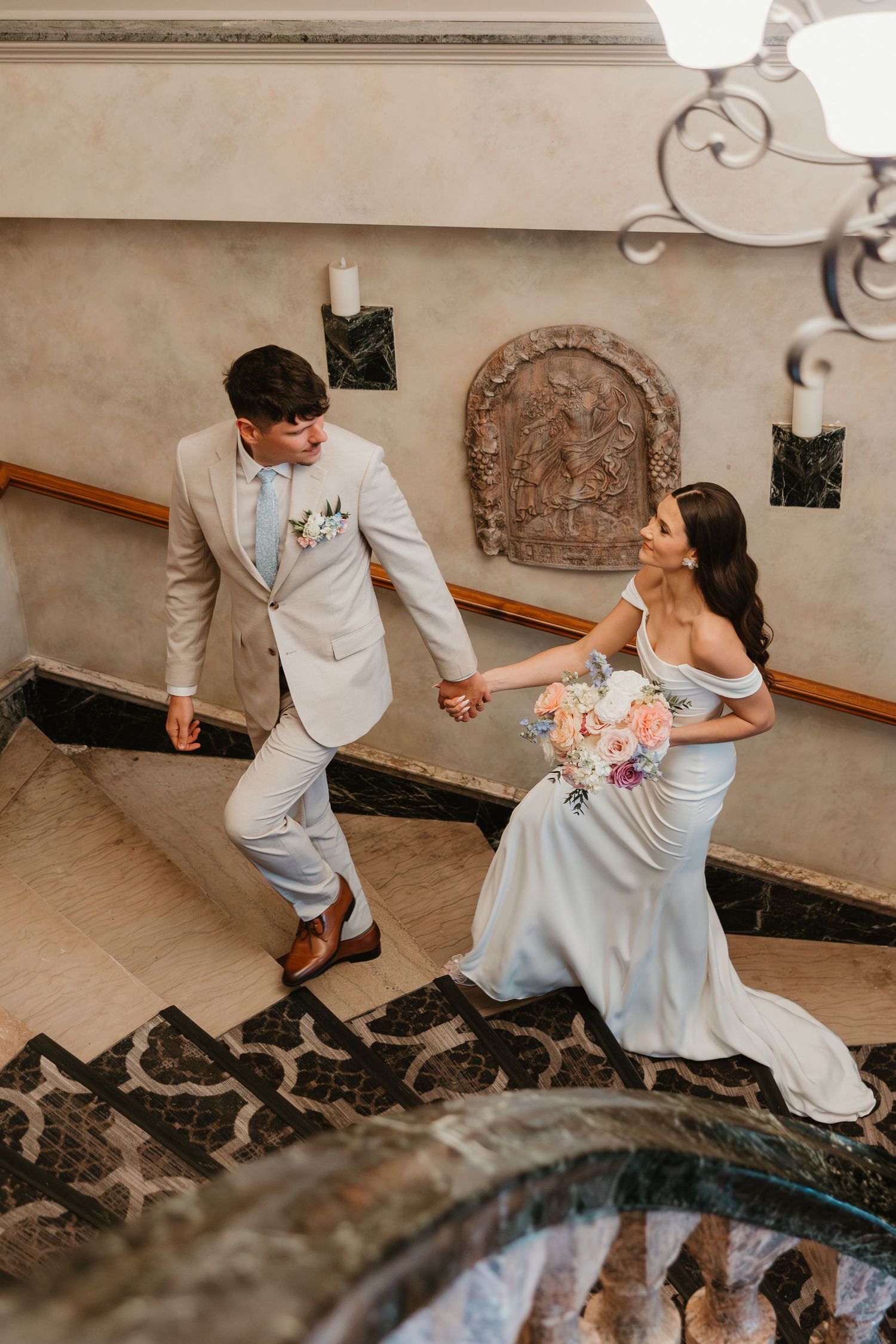 Bride and groom holding hands walking up elegant marble staircase in ornate venue.