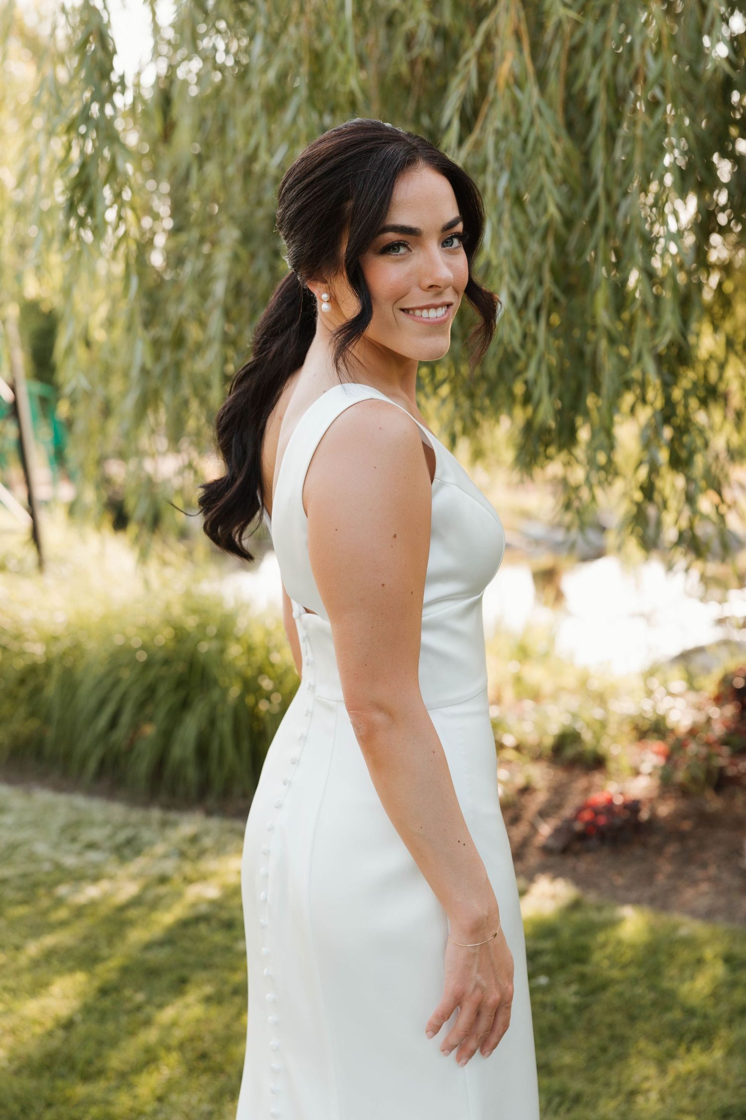 Smiling bride in sleek white gown posing outdoors near a weeping willow tree.