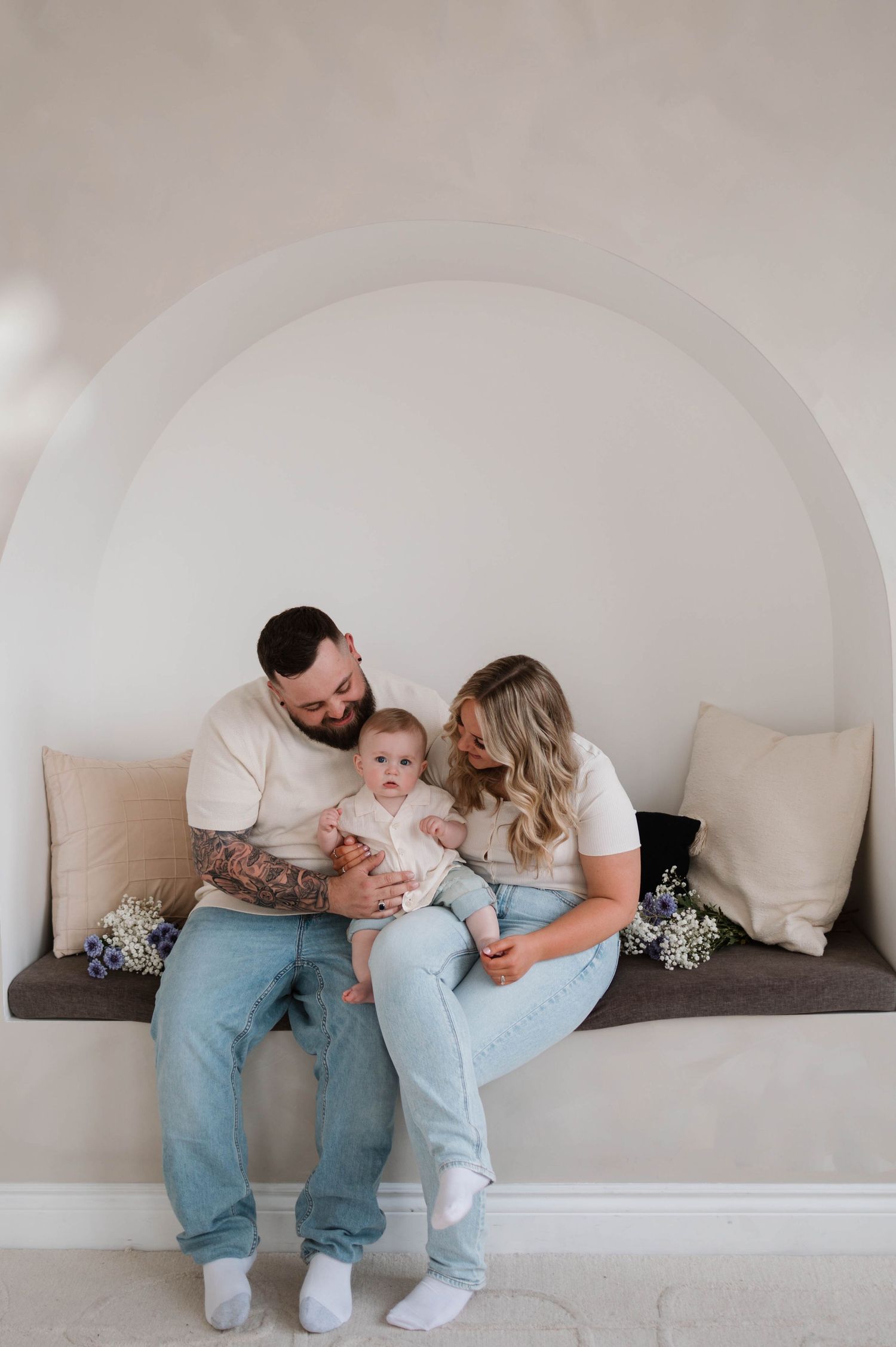 Family of three sitting in an arched alcove bench, parents smiling at their baby in a bright room.