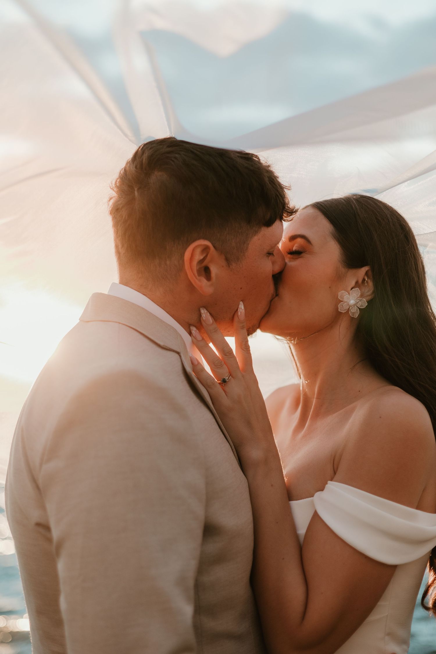 A bride and groom share a romantic kiss beneath a flowing veil at golden hour sunset.