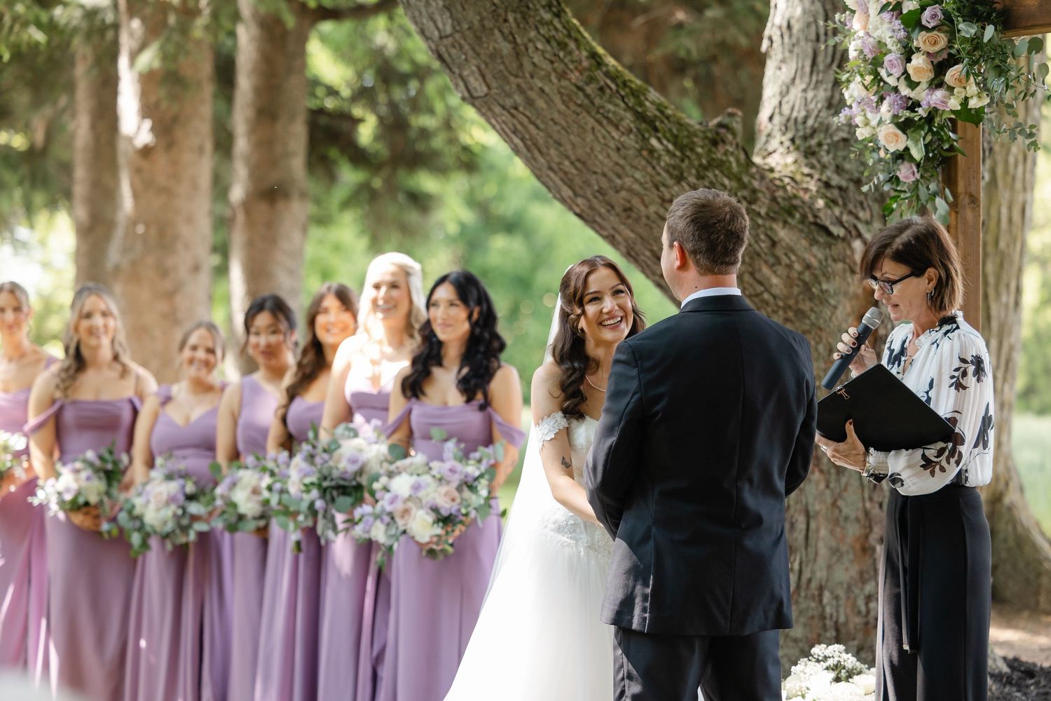 Bride and groom exchange vows outdoors as bridesmaids in lavender dresses hold matching floral bouquets.