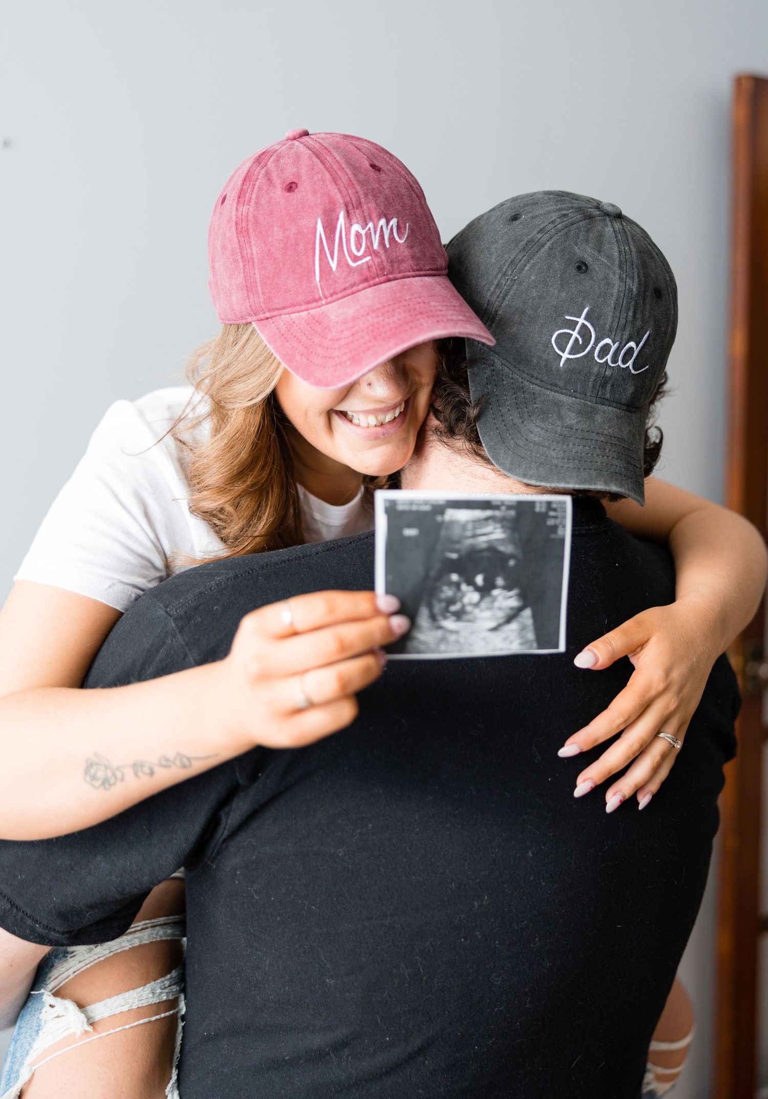 Couple hugging and smiling while holding ultrasound photo, wearing Mom and Dad baseball caps.